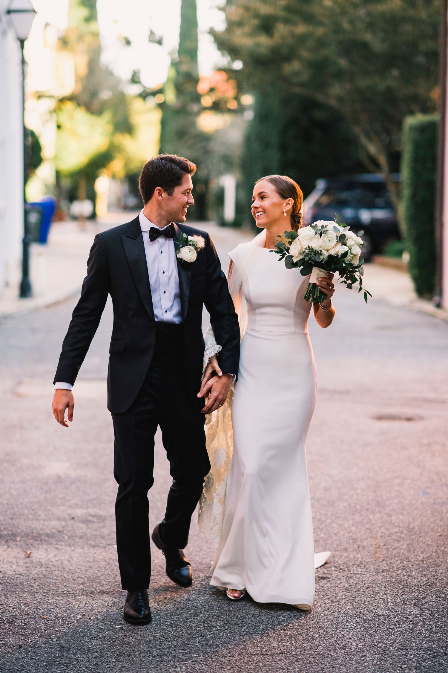 Romantic wedding couple walking and laughing together on a tree-lined path in formal attire.