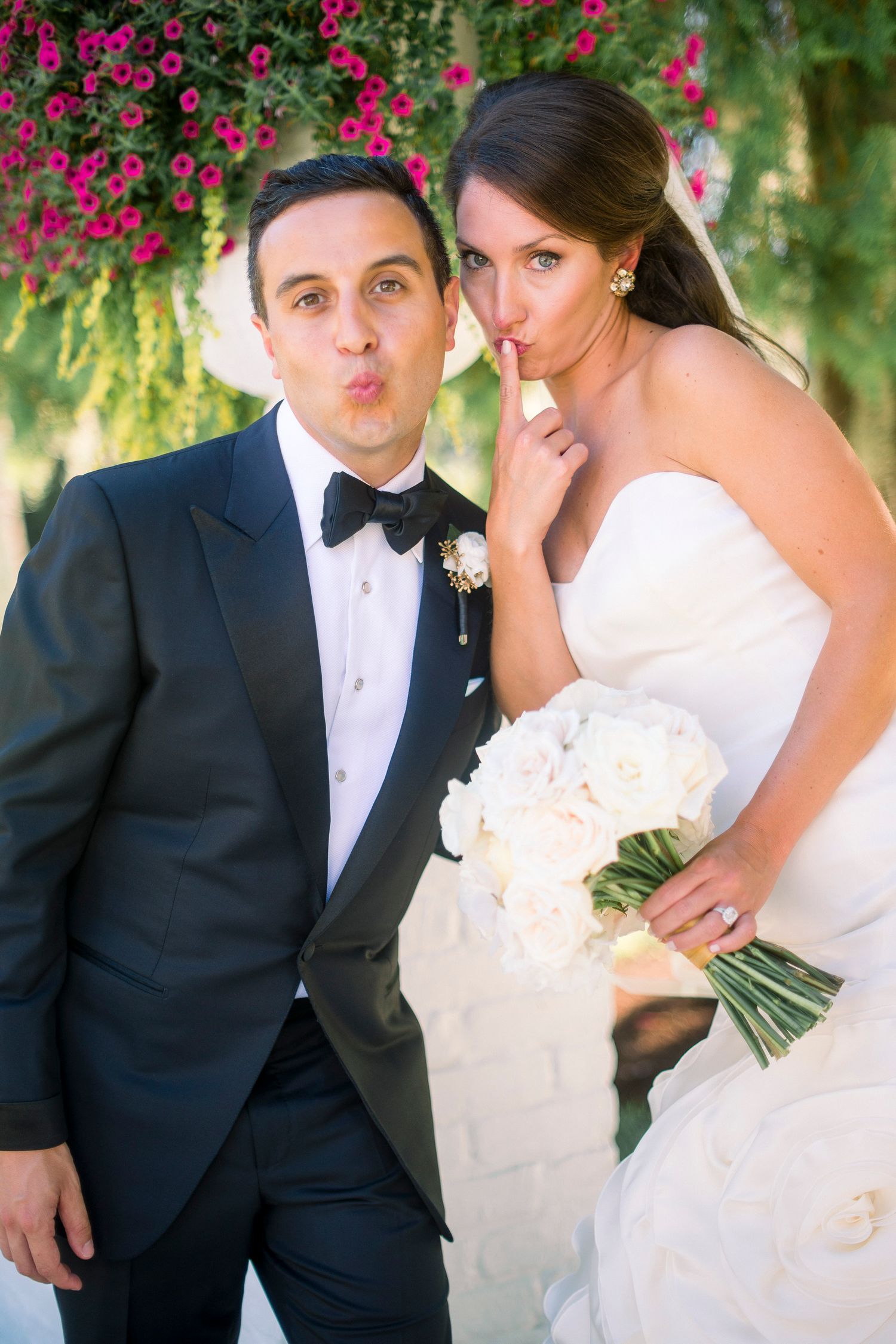 Romantic wedding couple poses among vibrant pink flowers in formal attire.