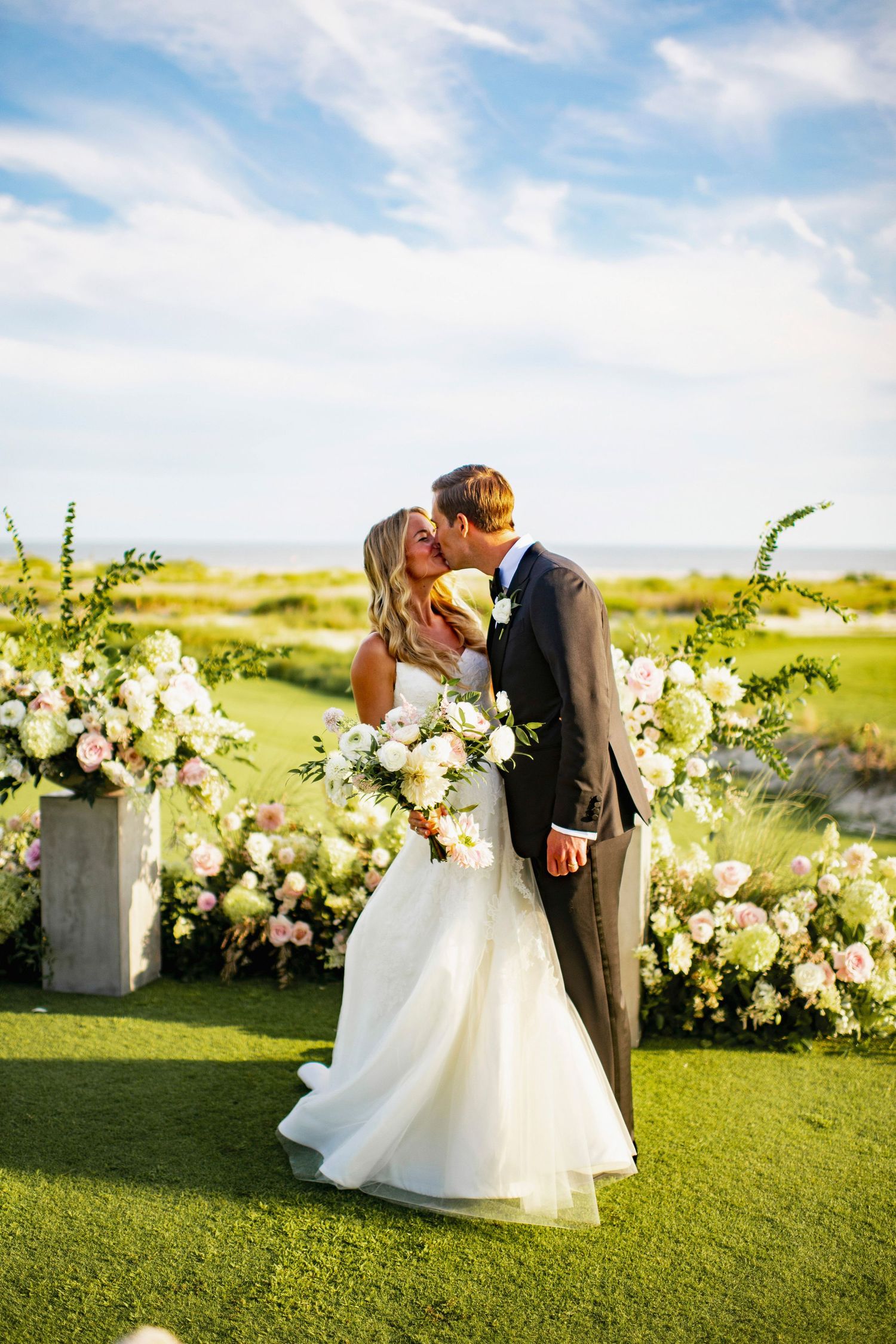 A romantic wedding scene with floral arrangements on a green lawn against a bright blue sky.