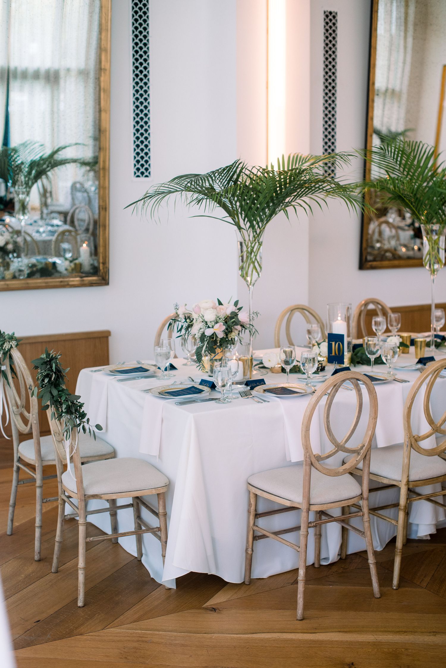 Elegant wedding reception table with white linens, gold chairs, palm fronds and floral centerpieces in a bright dining room.