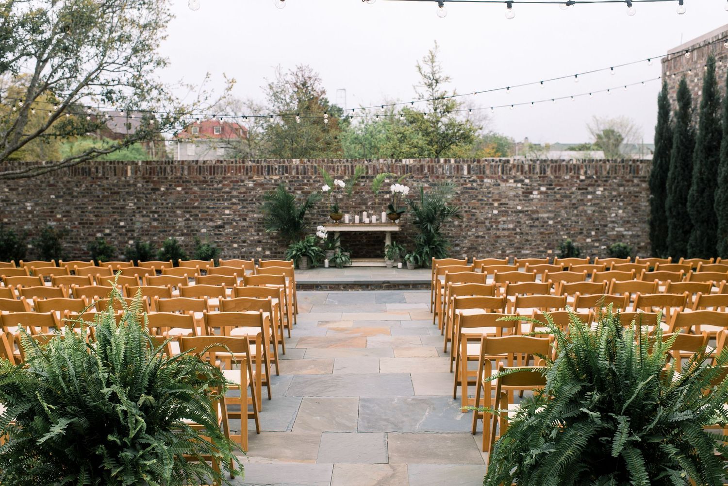 Rustic outdoor wedding venue with wooden chairs arranged in rows and string lights hanging overhead.