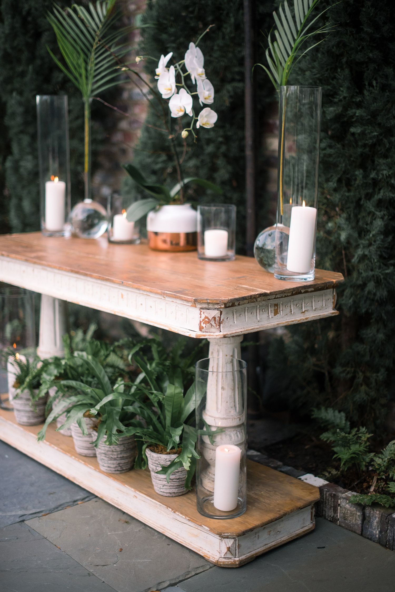 Rustic wooden console table decorated with white orchids, pillar candles, and potted ferns against a dark garden backdrop.