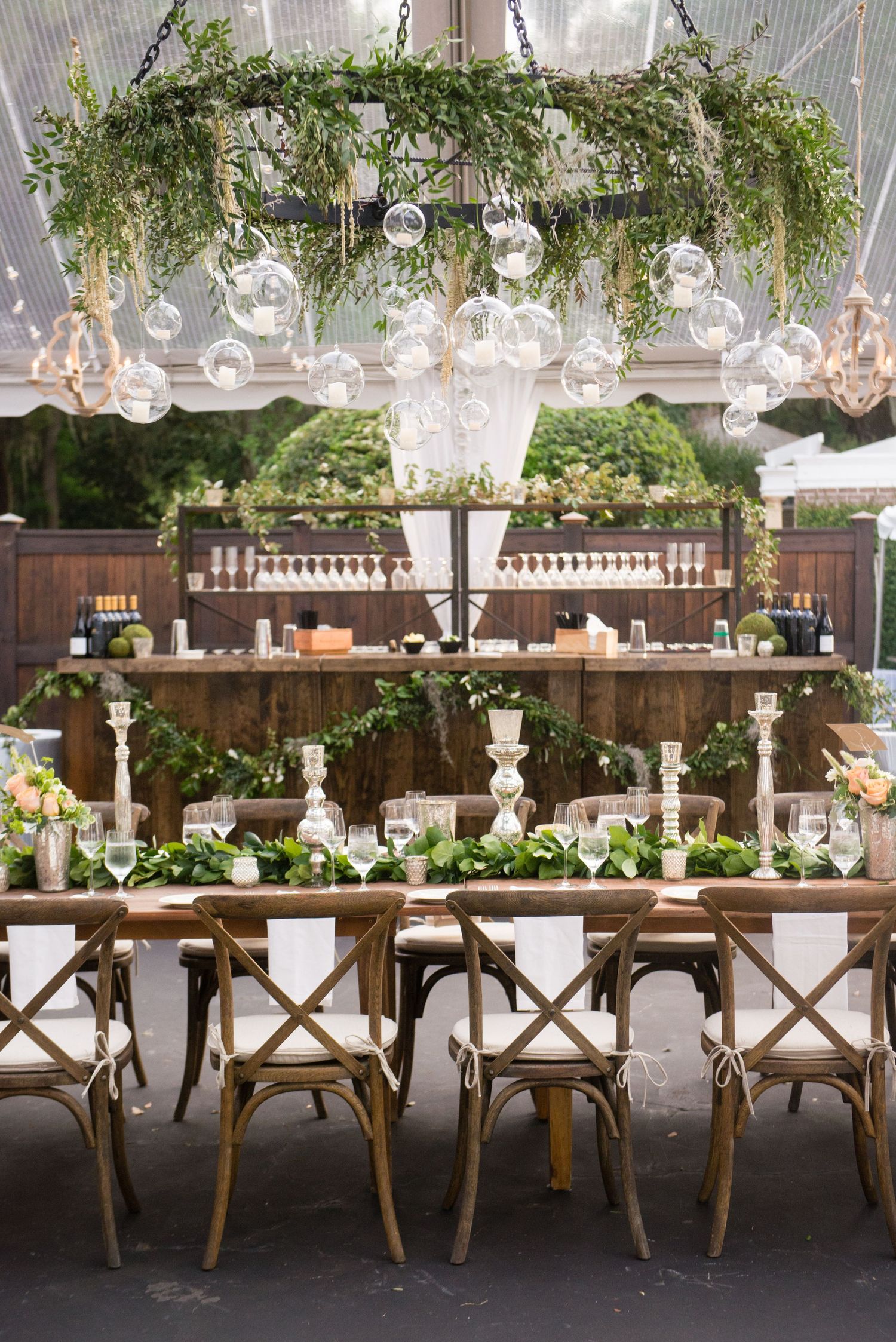 Elegant outdoor wedding reception table with rustic wooden chairs and hanging crystal chandeliers under a white tent.