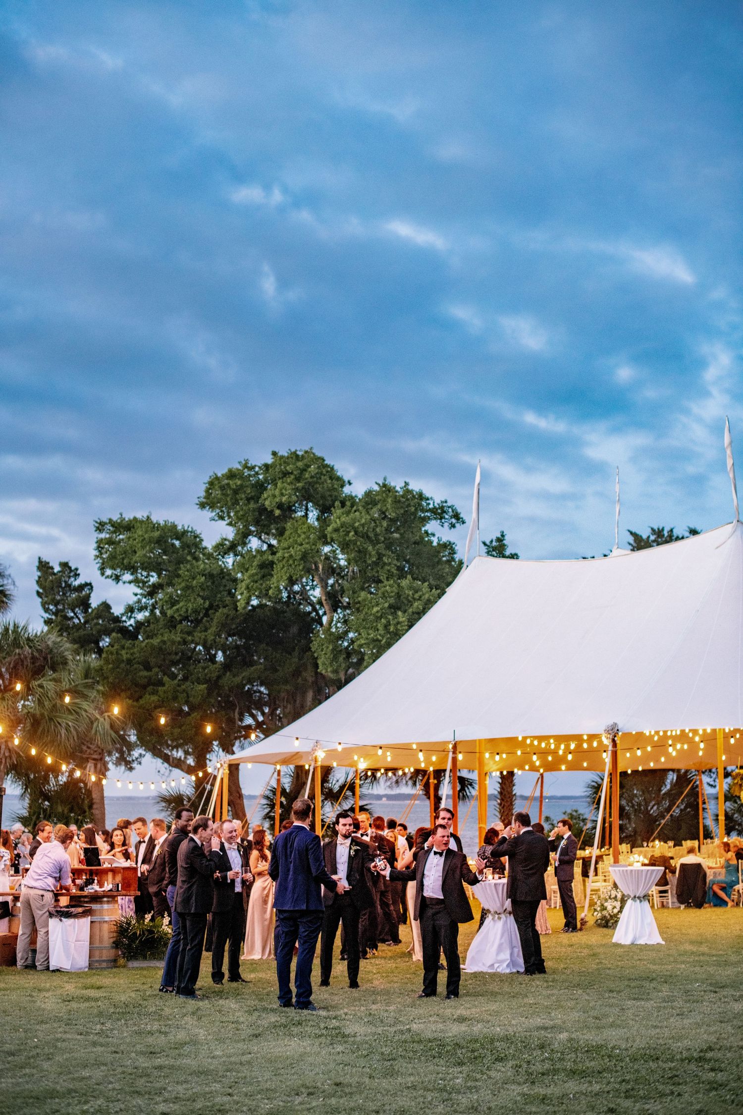 Wedding guests gather under illuminated white tents at dusk during an elegant outdoor reception celebration.