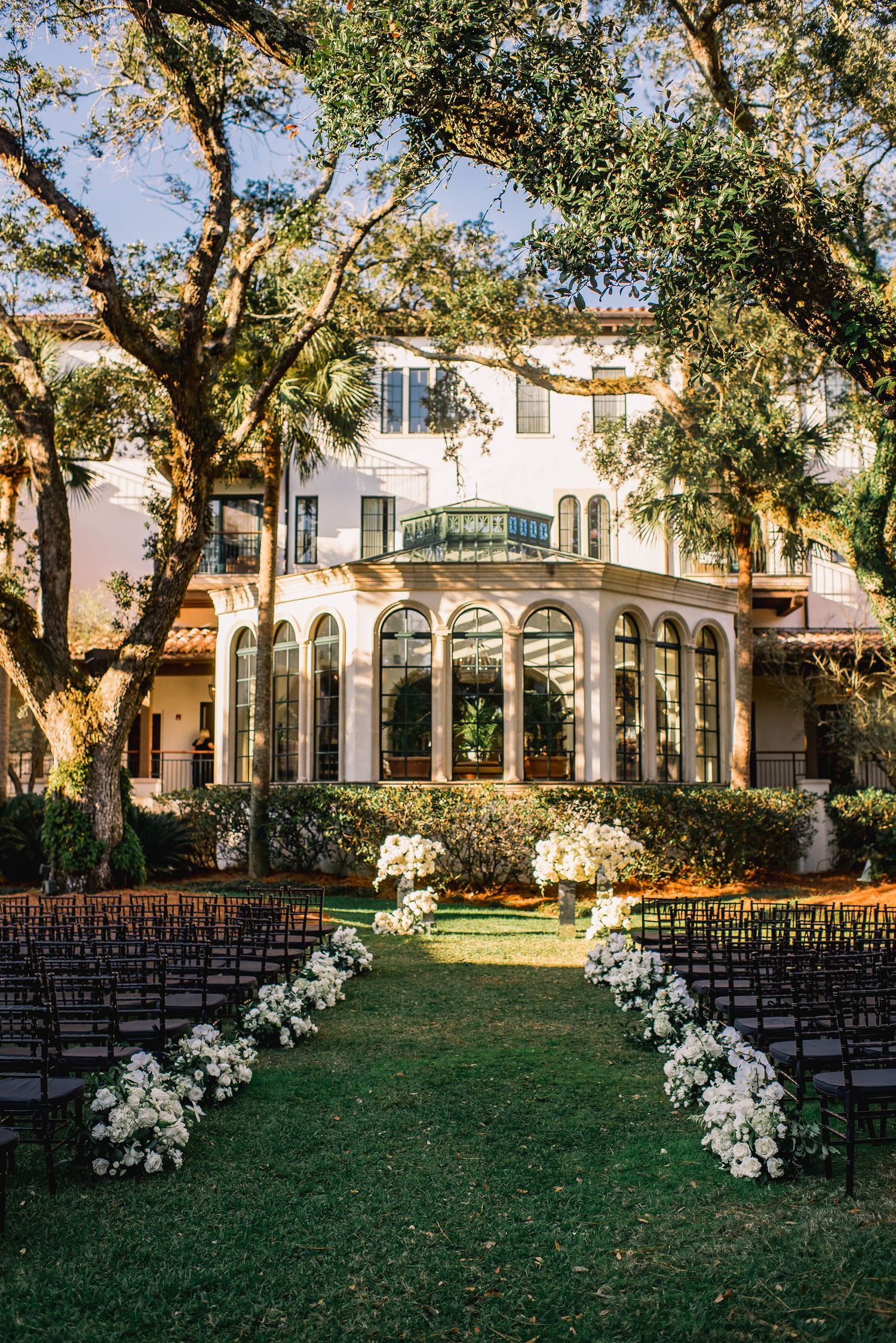 Elegant outdoor wedding ceremony setup with white floral arrangements and dark chairs beneath towering trees at a Spanish-style venue.