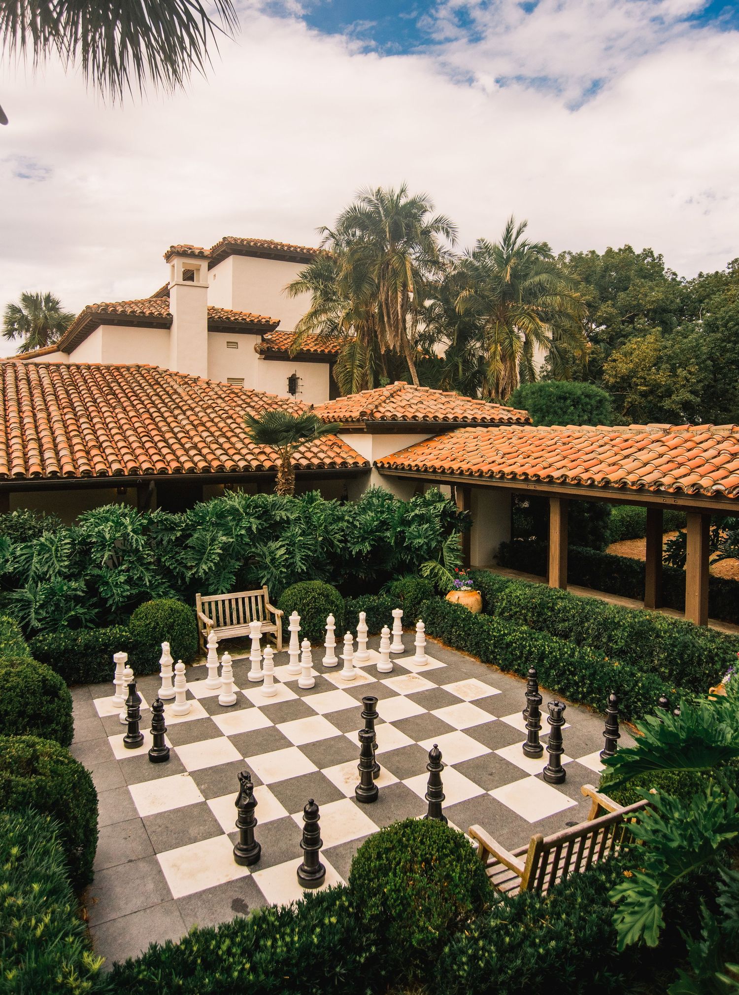 Large outdoor chess set with black and white pieces on a patio surrounded by lush tropical landscaping and Spanish tile roof.