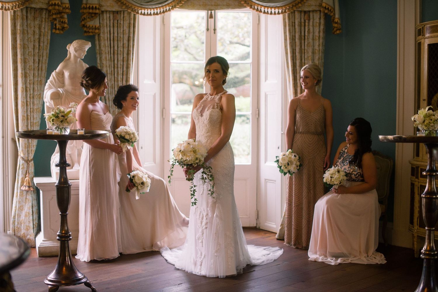 Wedding party in soft pink dresses gathers near sunlit windows in an elegant room during preparation.