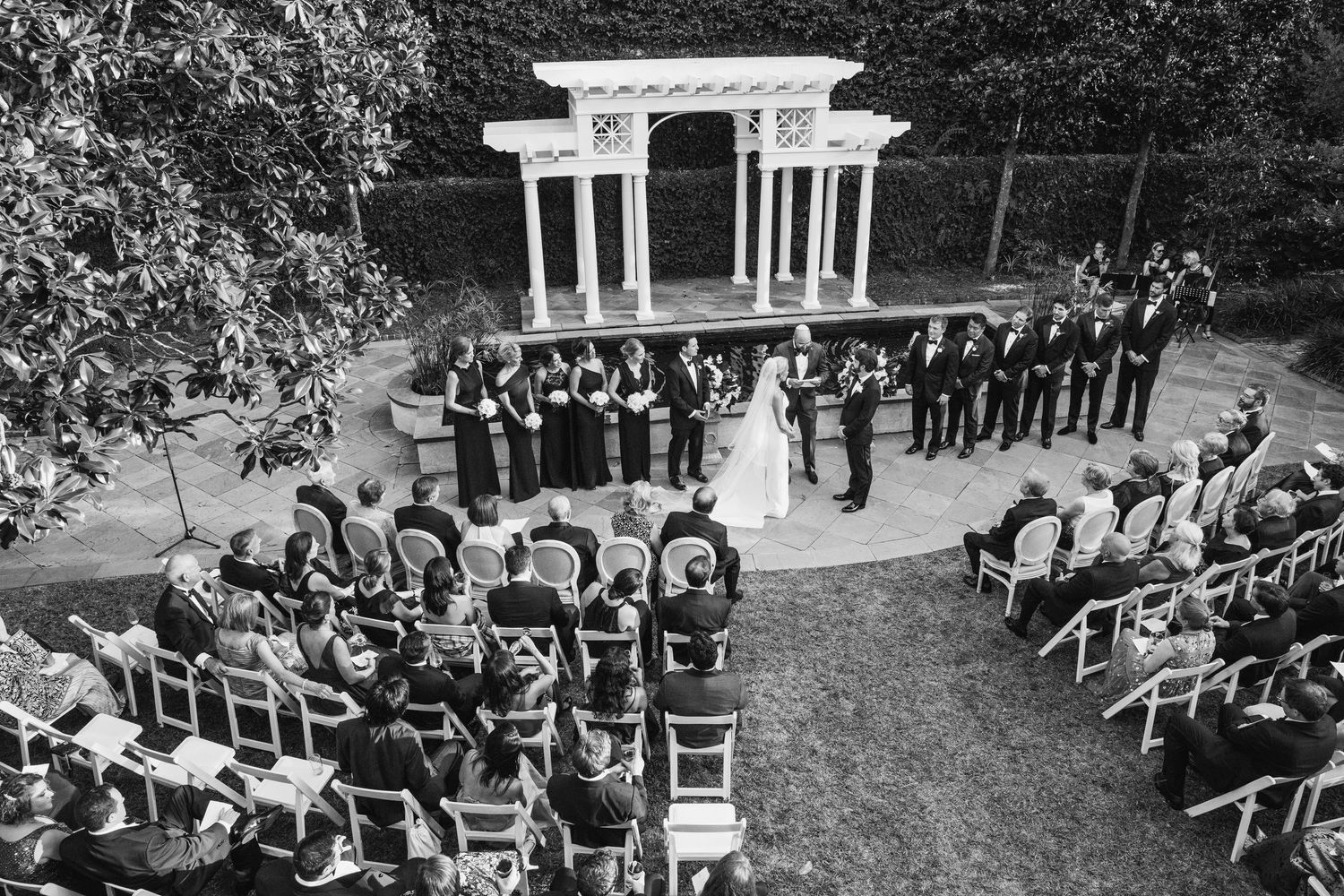A black and white panoramic photo shows guests seated at an outdoor garden wedding ceremony with white chairs.