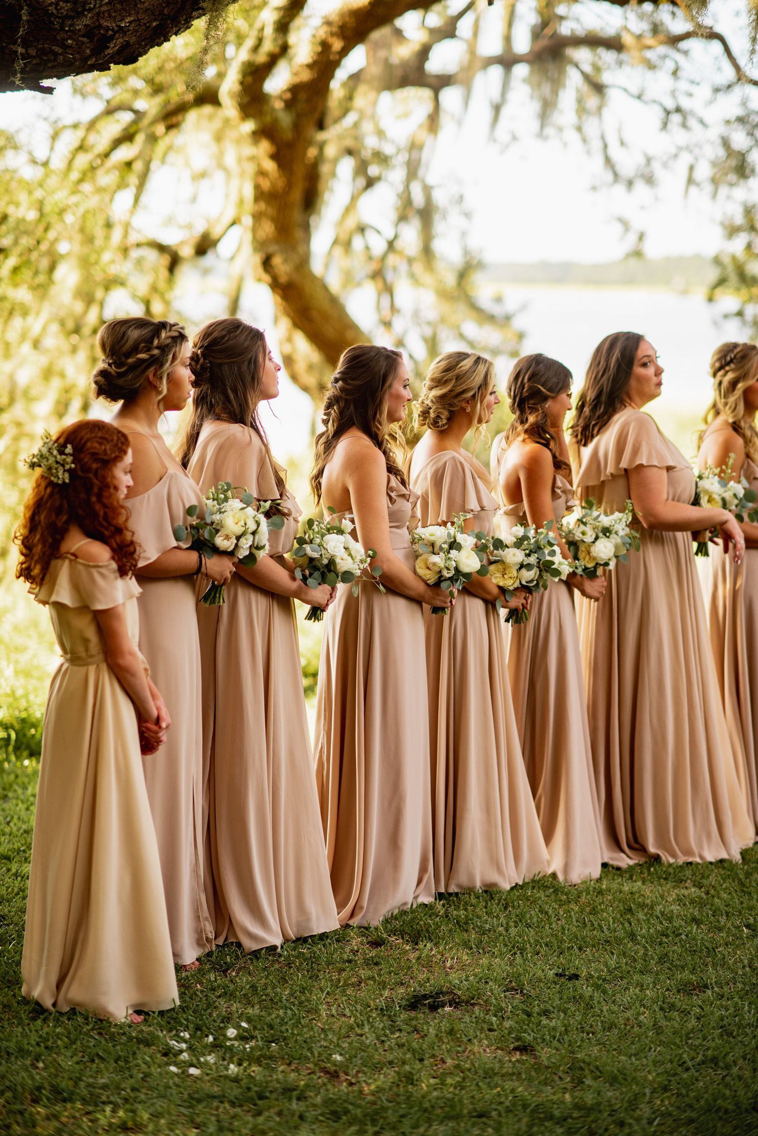 A row of bridesmaids in champagne-colored dresses stand in line holding bouquets on a grassy hill.
