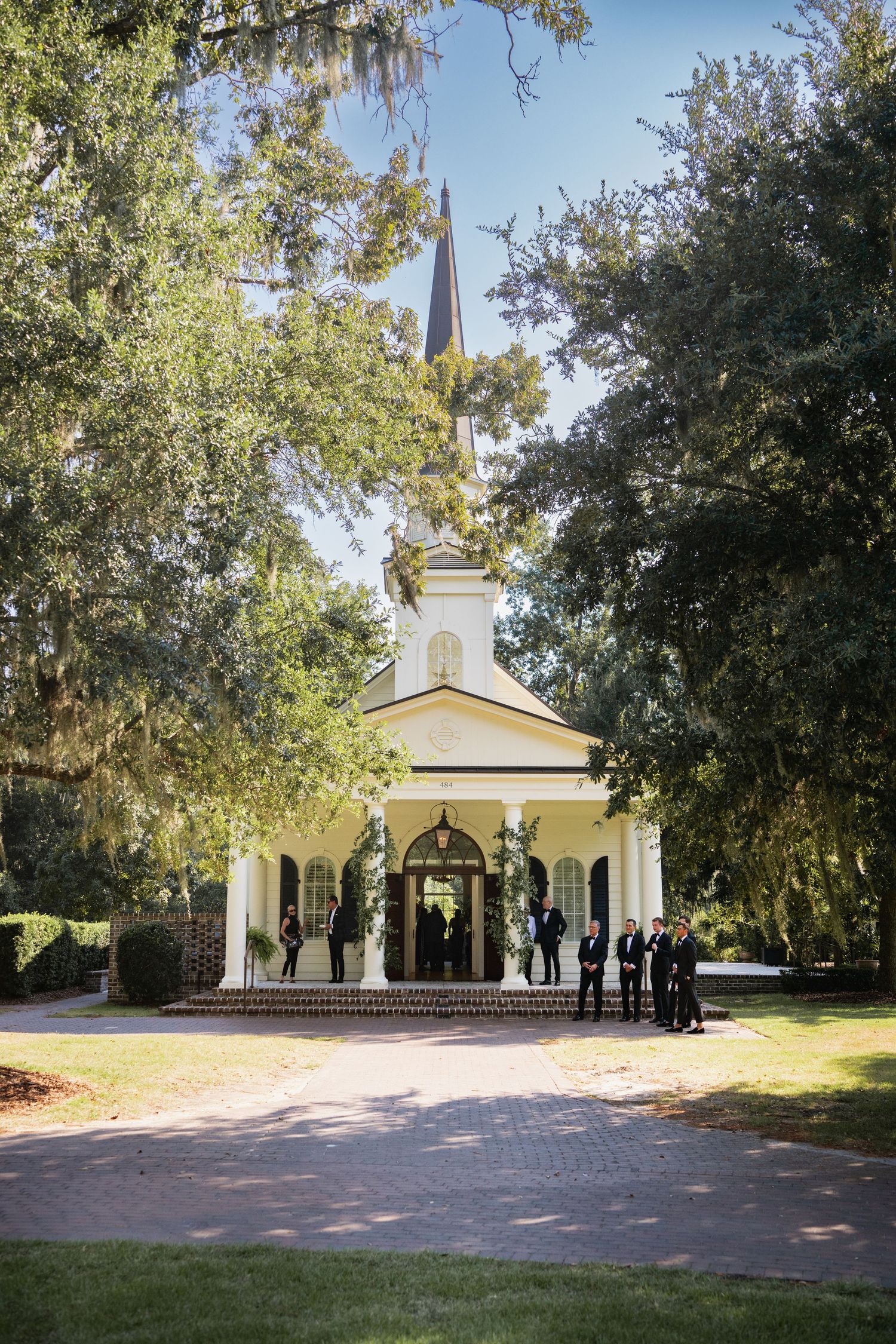 A charming white chapel with tall spire stands framed by Spanish moss-draped trees on a brick pathway.