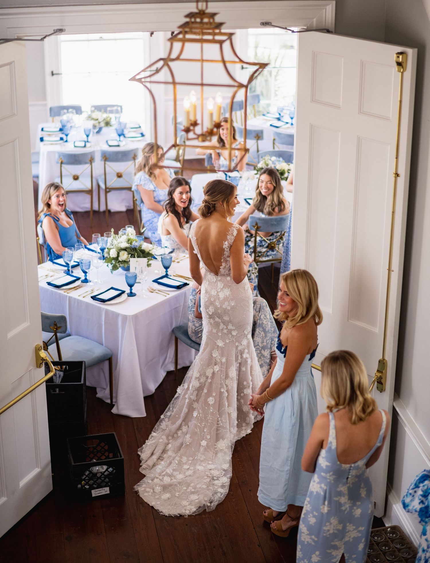 A bride in a lace wedding dress gets ready in a bright room decorated with elegant chandeliers and table settings.
