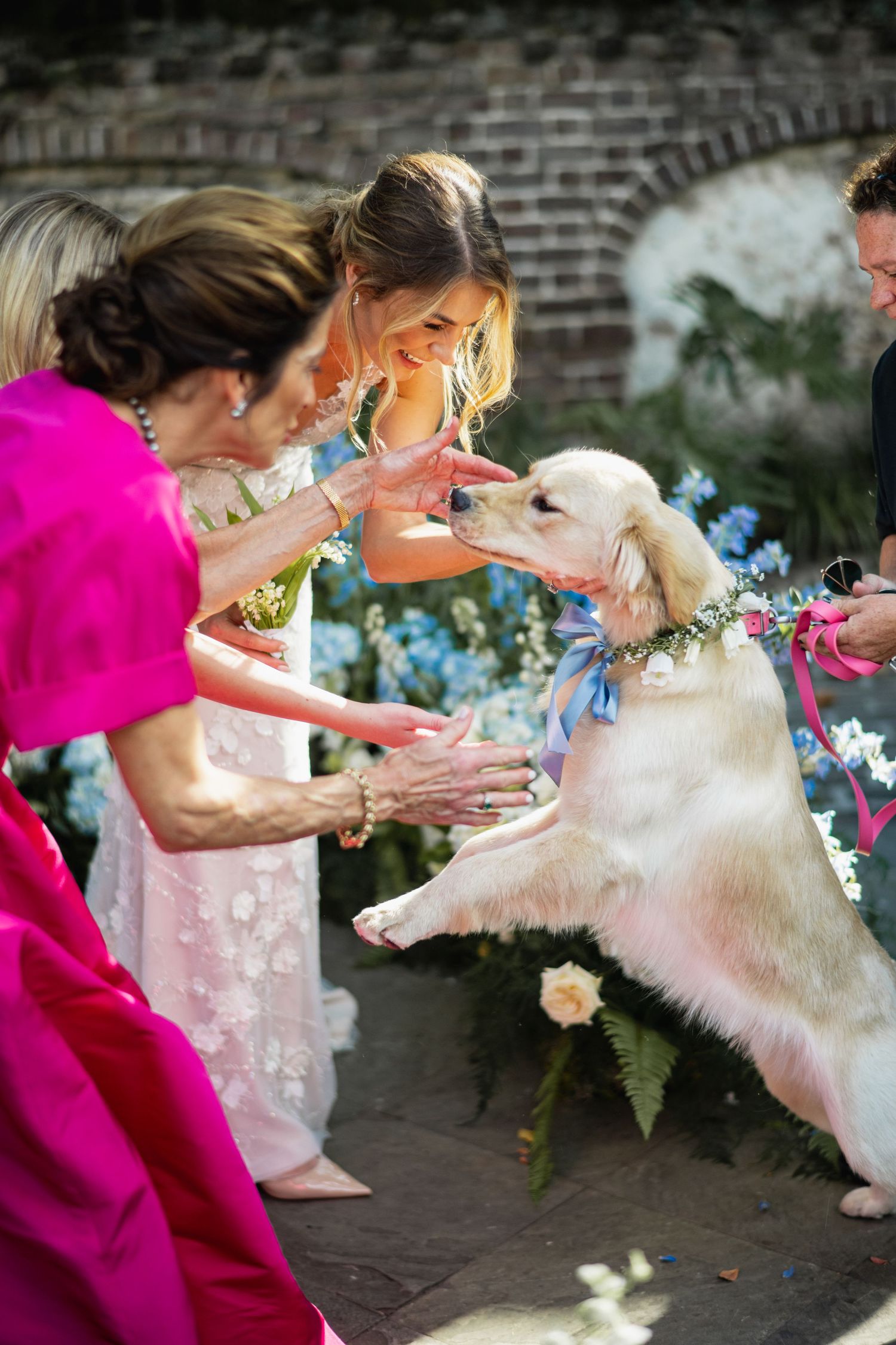 Wedding party members in pink dresses help adjust a bride's flowing white dress on a brick patio.