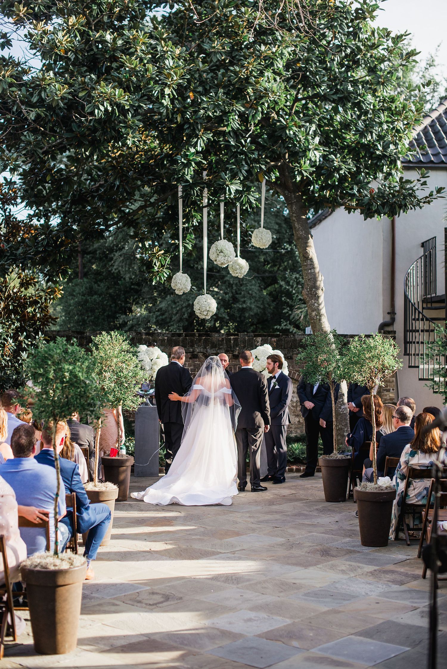 A wedding ceremony takes place in a courtyard with white lanterns hanging from trees and guests gathered around.