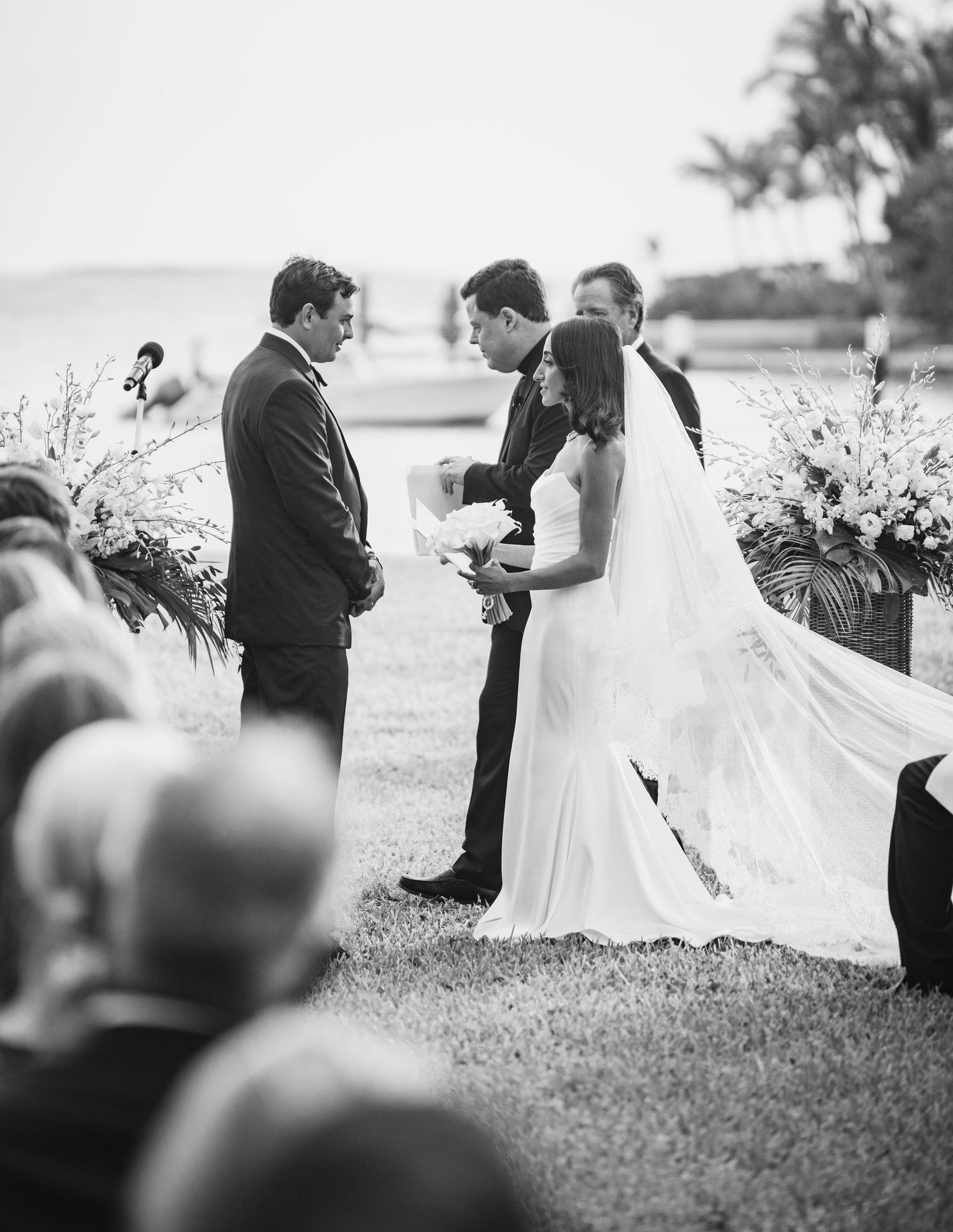 Black and white photograph of an outdoor wedding ceremony by the water with floral arrangements and a flowing bridal veil.