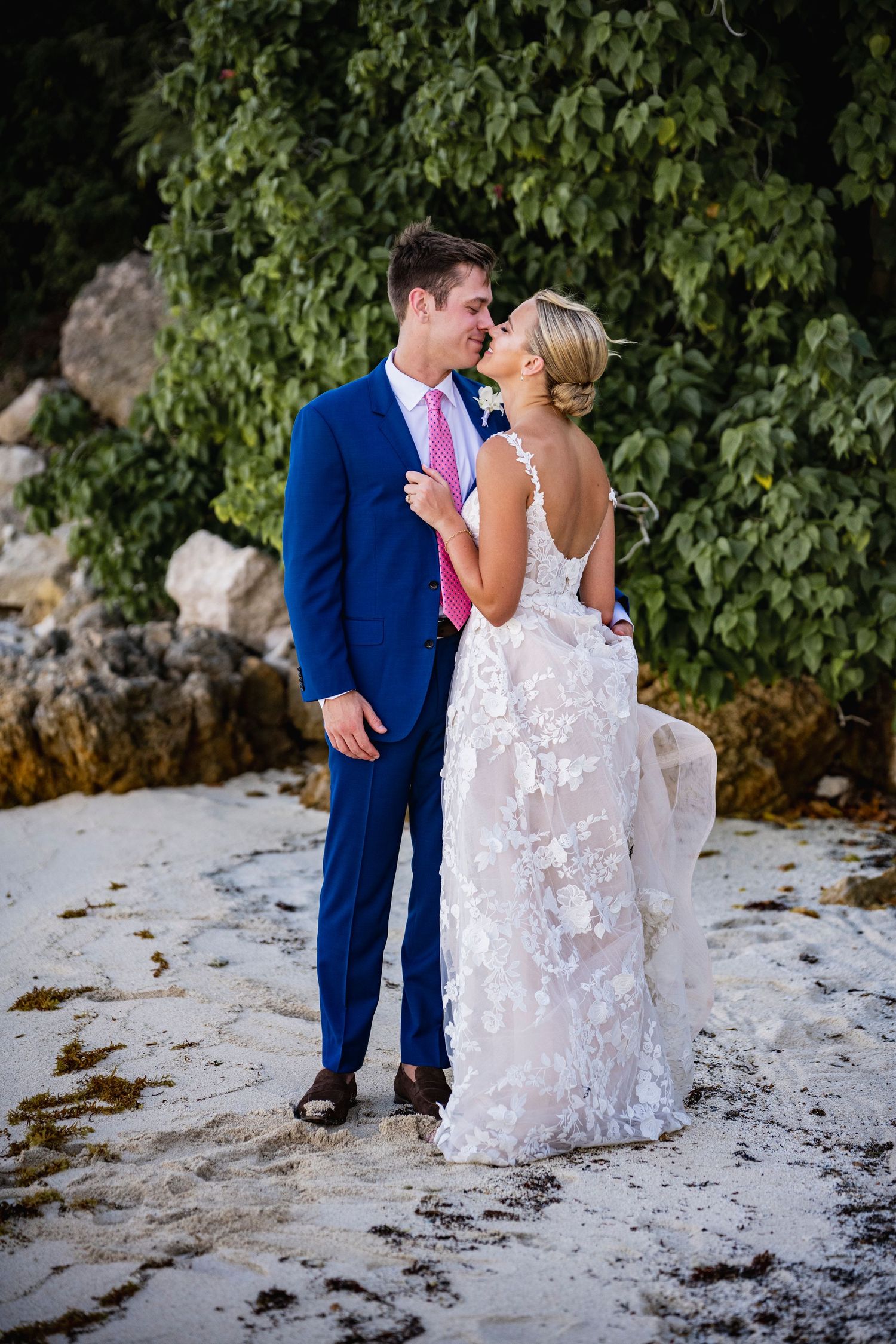 A couple shares an intimate moment on a beach, wearing a blue suit and white lace wedding dress.