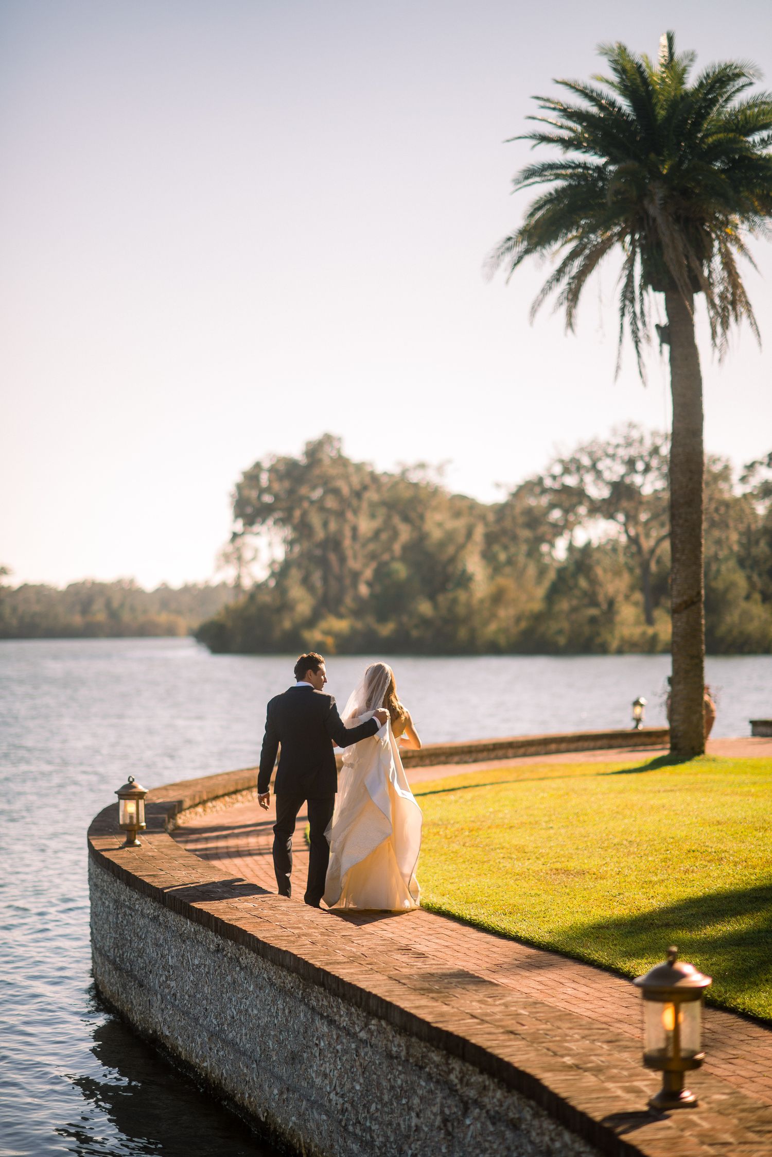 A couple stands on a stone wall overlooking a lake at sunset with palm trees and lanterns creating a romantic scene.