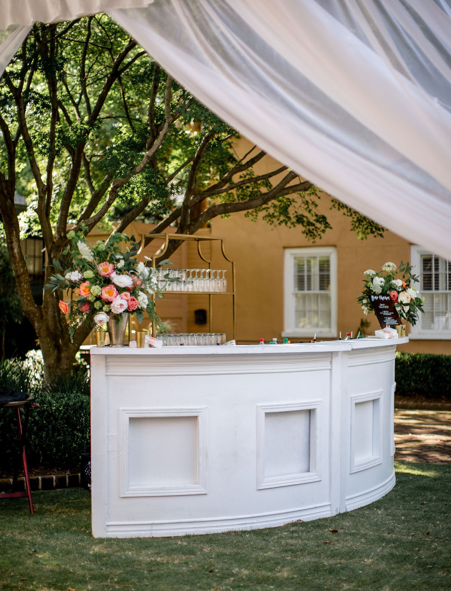 Elegant curved white outdoor bar setup with draped fabric and floral arrangements at a garden event venue.