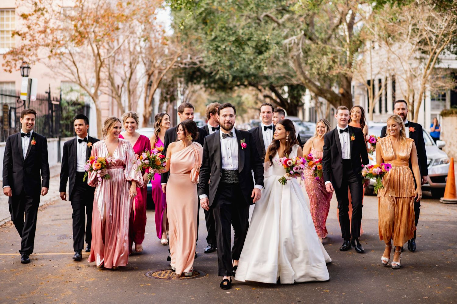 Wedding party in formal attire walks together down tree-lined path during autumn season.