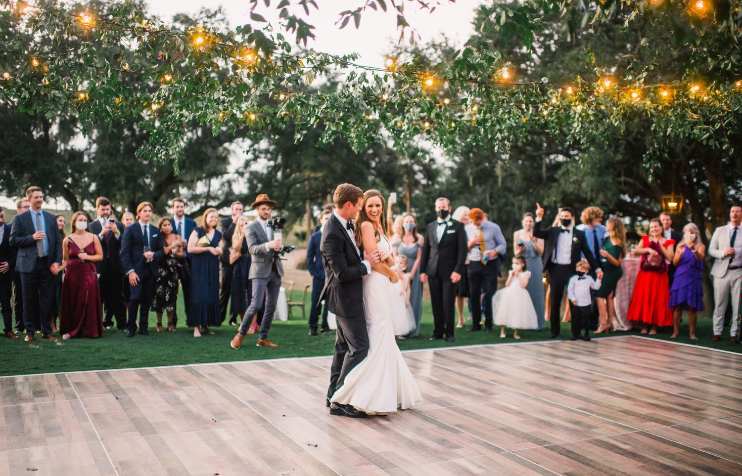 Wedding guests gather around an outdoor dance floor at sunset while a couple shares their first dance together.
