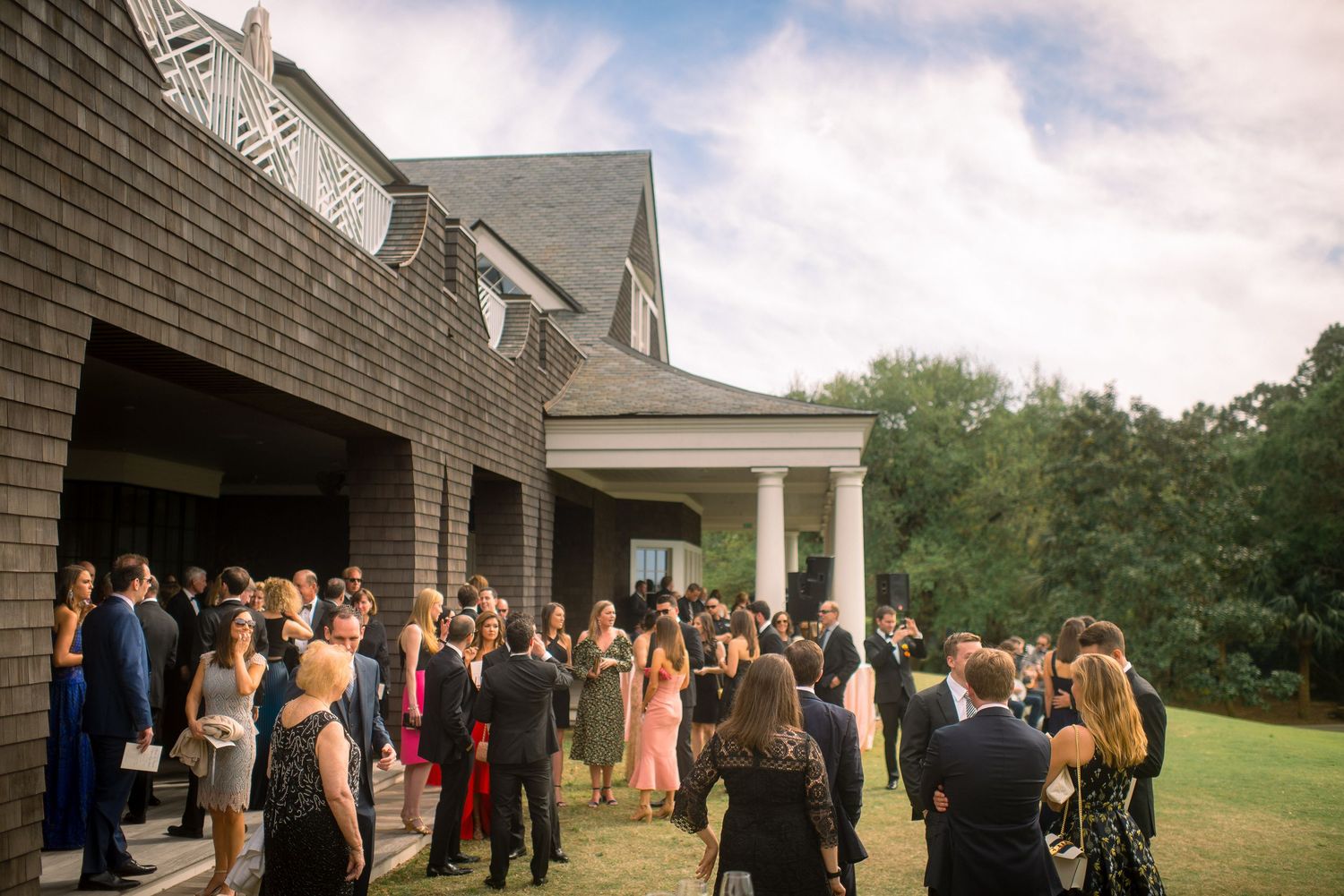 A large group of wedding guests gather outside a gray shingled building with white columns on a cloudy day.