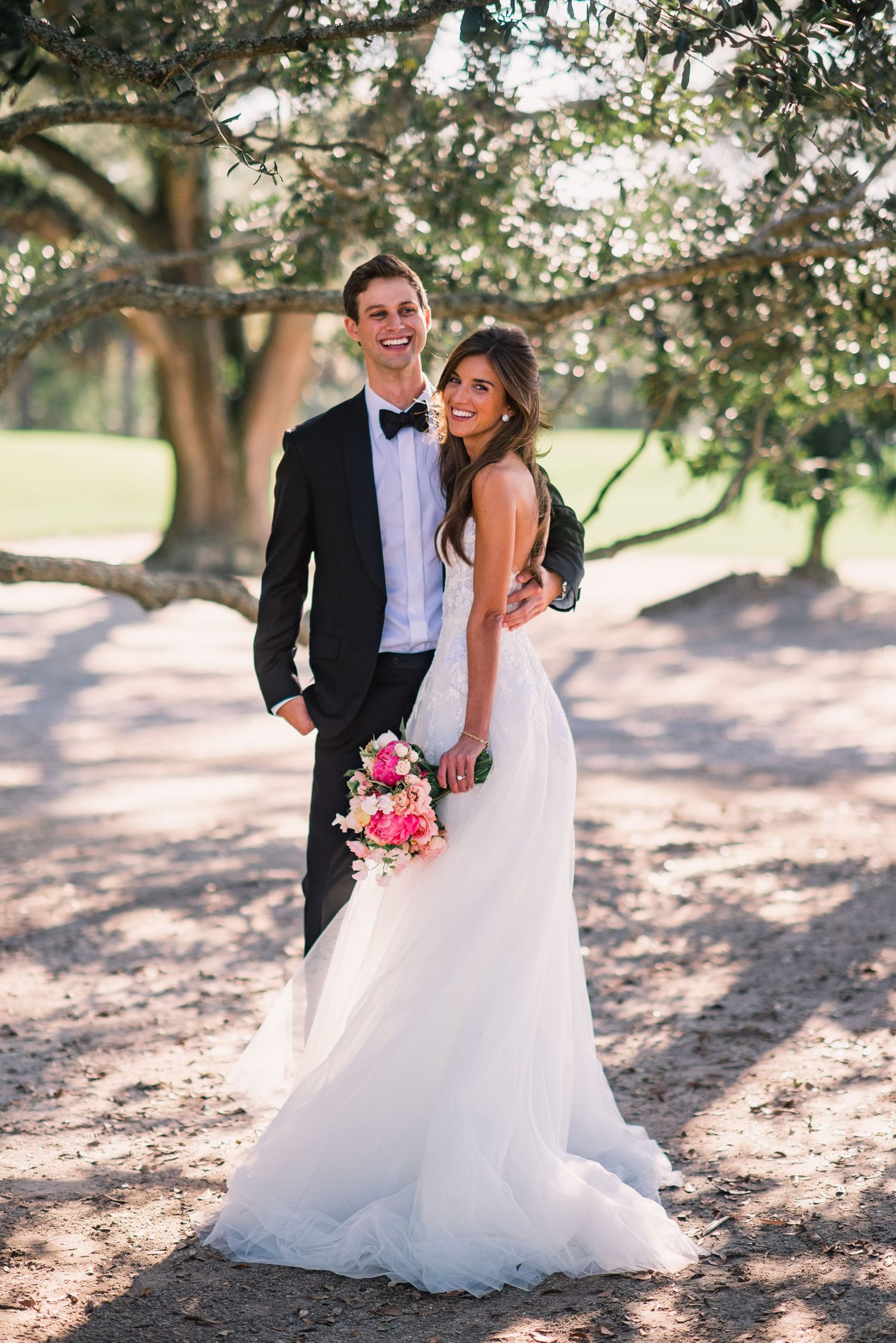 A couple in formal wedding attire poses under a tree with dappled sunlight, the bride holding a pink bouquet.