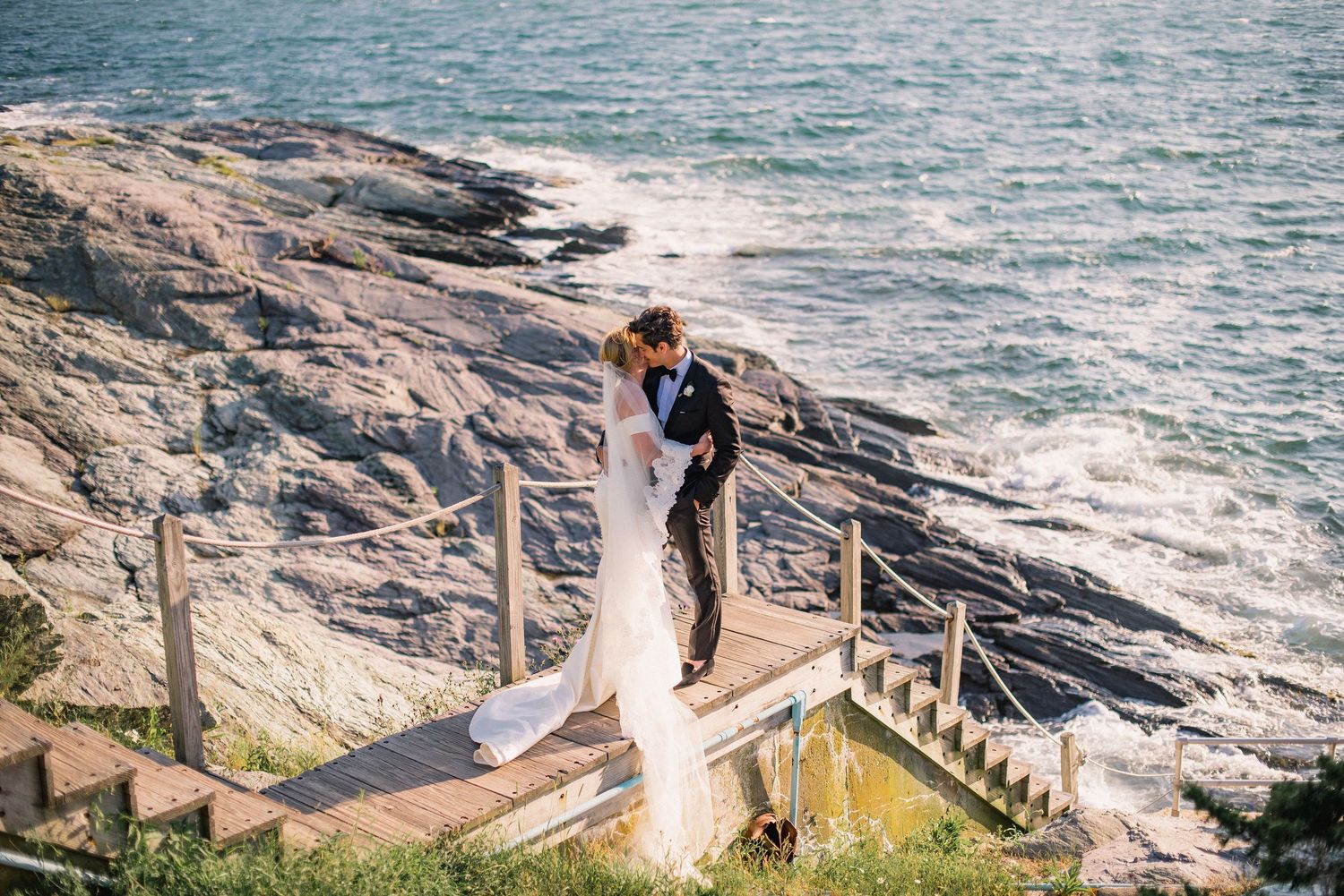 A couple poses along a scenic coastal cliff with crashing waves and wooden railings during a romantic photoshoot.
