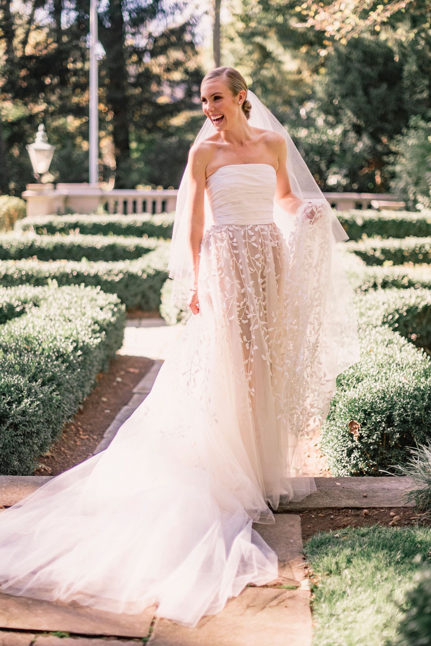 A bride in a strapless lace gown poses in a formal garden setting with manicured hedges.