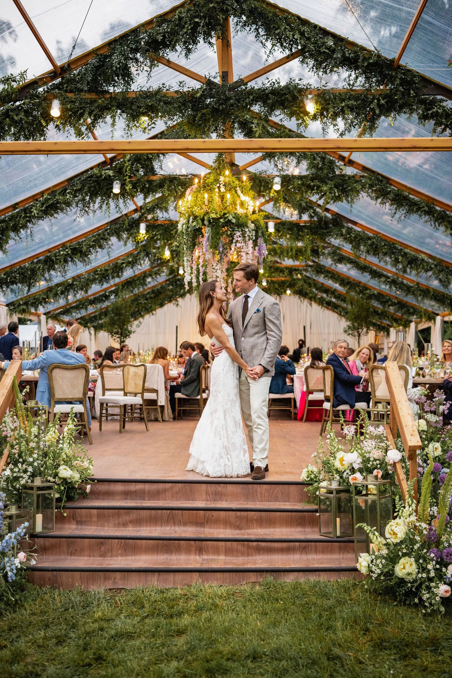 An elegant wedding reception setup in a clear tent with greenery and string lights.