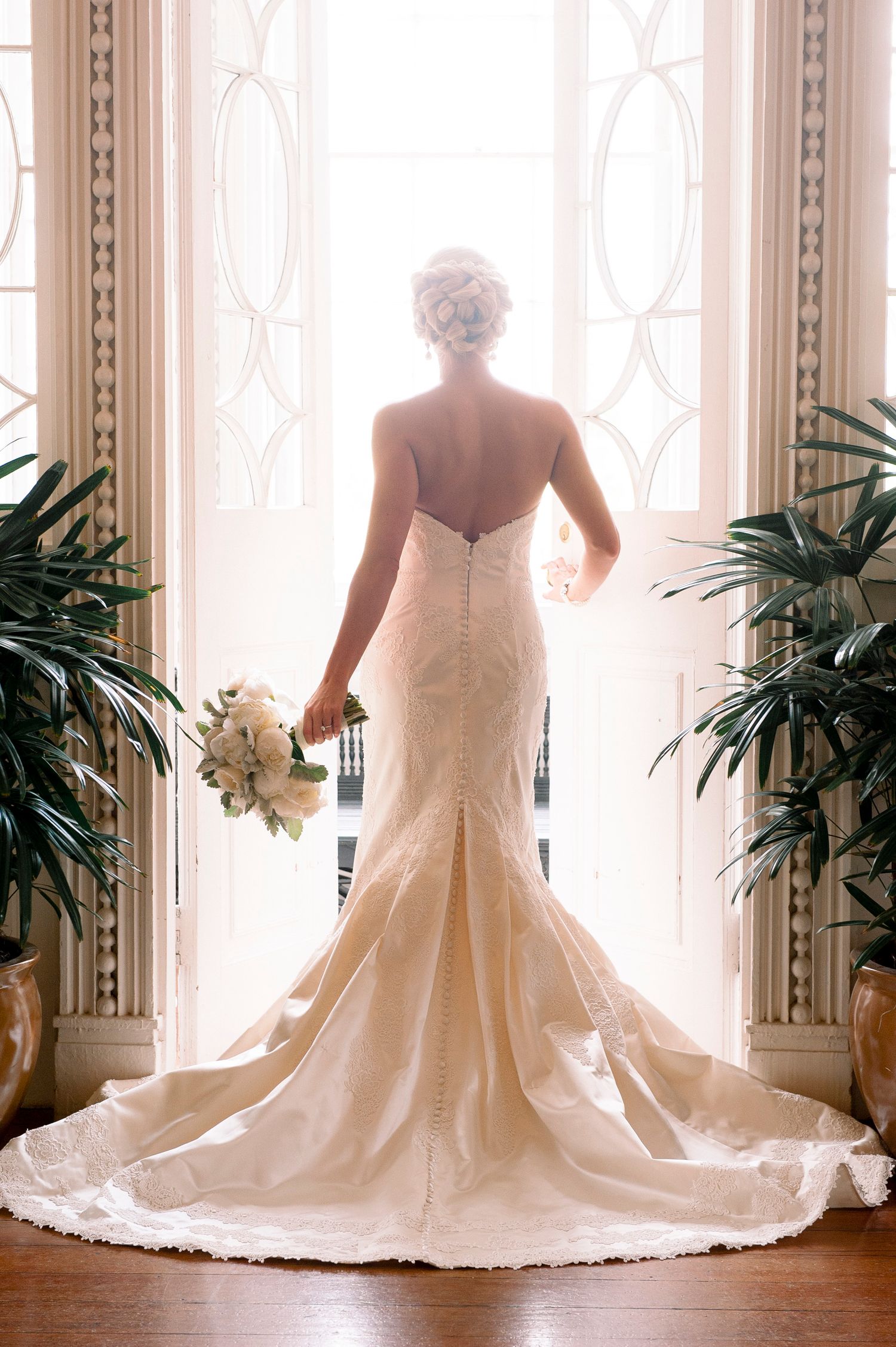 Elegant mermaid wedding dress with long train photographed from behind against bright windows with potted palms.