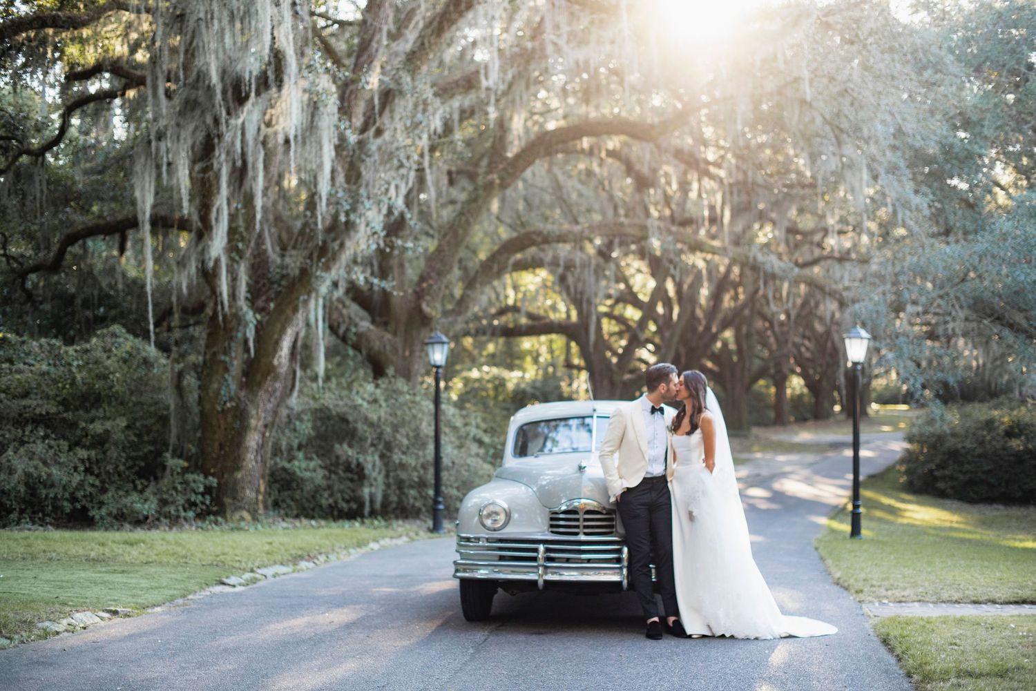 A couple shares a romantic moment under Spanish moss draped oak trees in a sunlit garden setting.