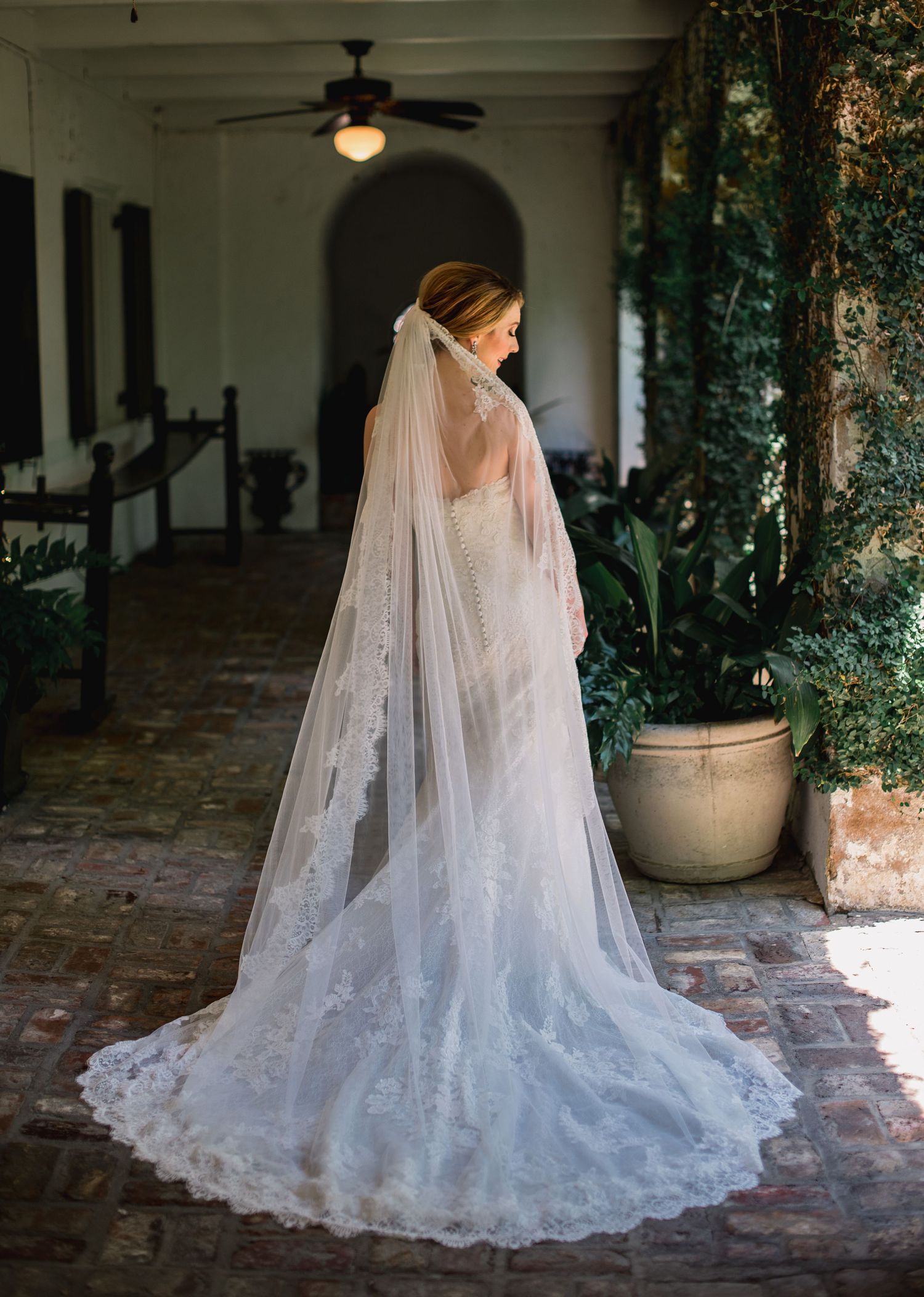 A bride in a flowing white lace wedding gown and cathedral veil poses on a covered porch with stone flooring.