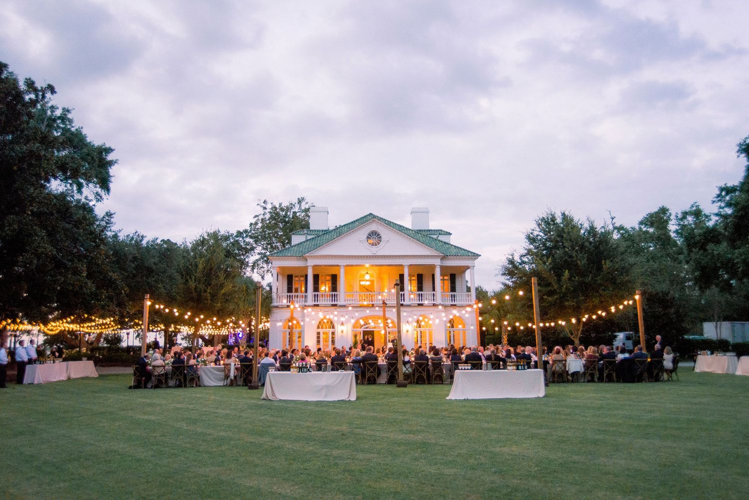 Panoramic view of an elegant white mansion wedding venue illuminated at dusk with string lights and guests on the lawn.