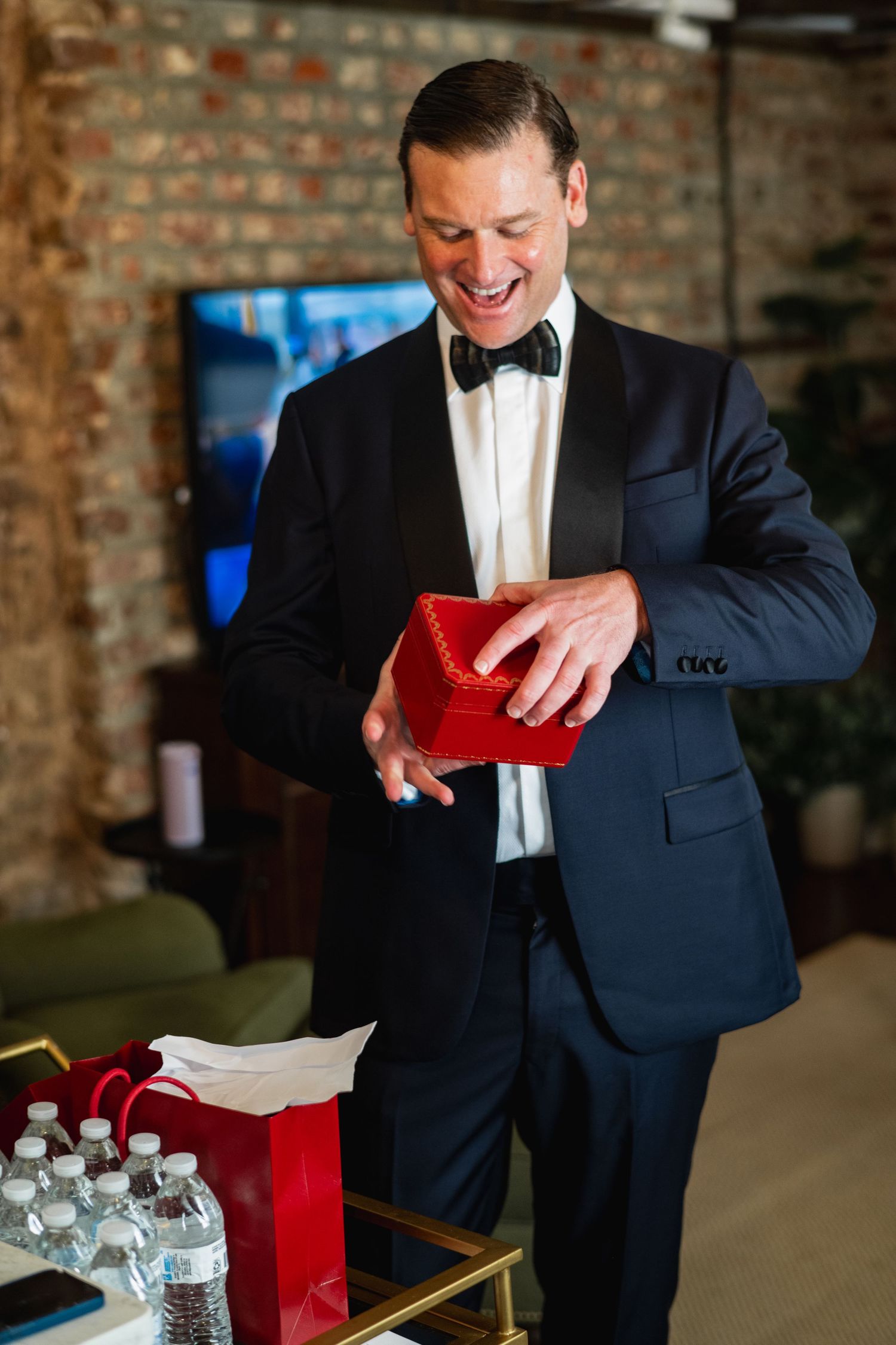 Person in formal attire with bow tie opens red gift bag while standing against exposed brick wall background.