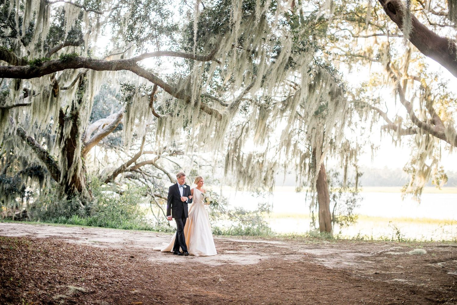Wedding couple stands beneath majestic oak trees draped with Spanish moss during golden hour.