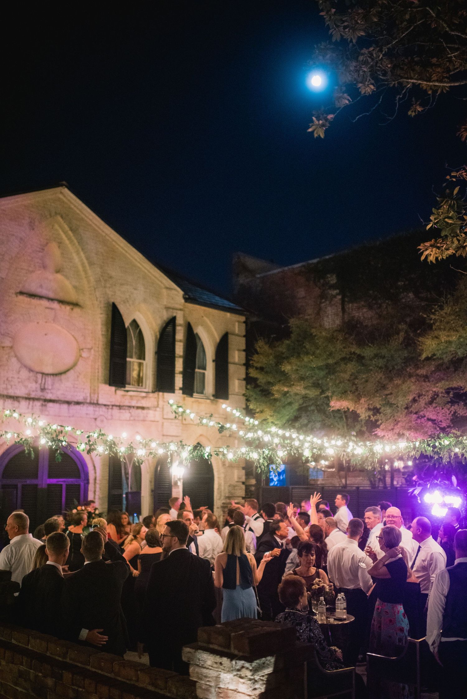 Outdoor evening wedding reception with string lights illuminating guests dancing and mingling under a moonlit sky.