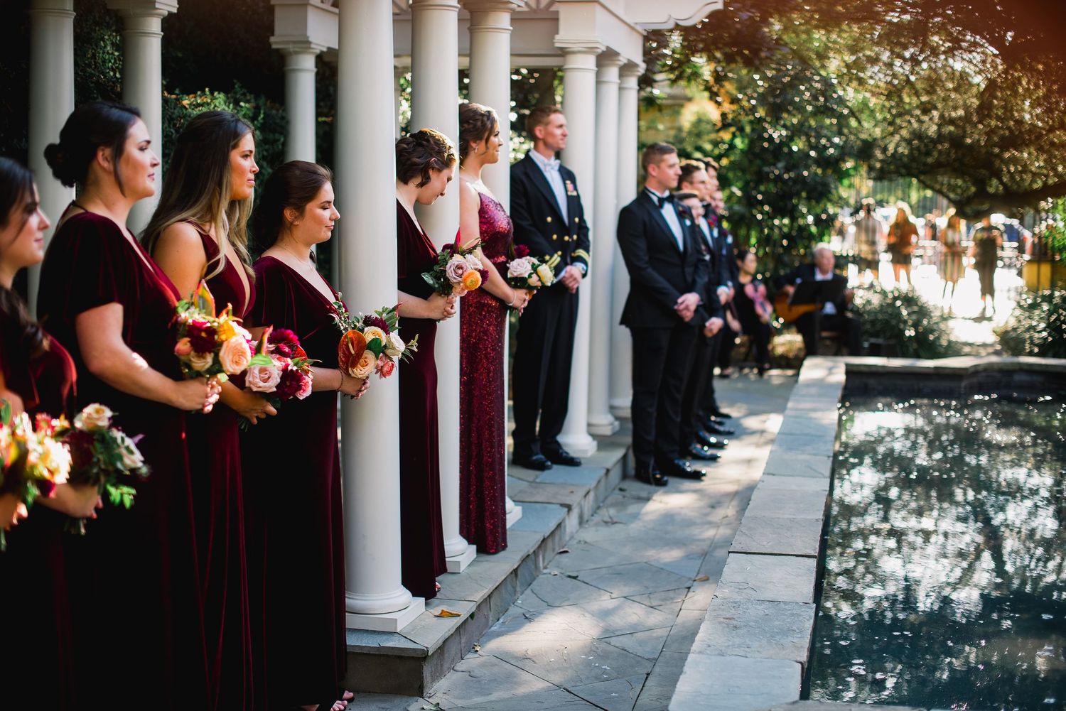 Wedding party stands in line along white columns during an outdoor ceremony with burgundy and black attire.