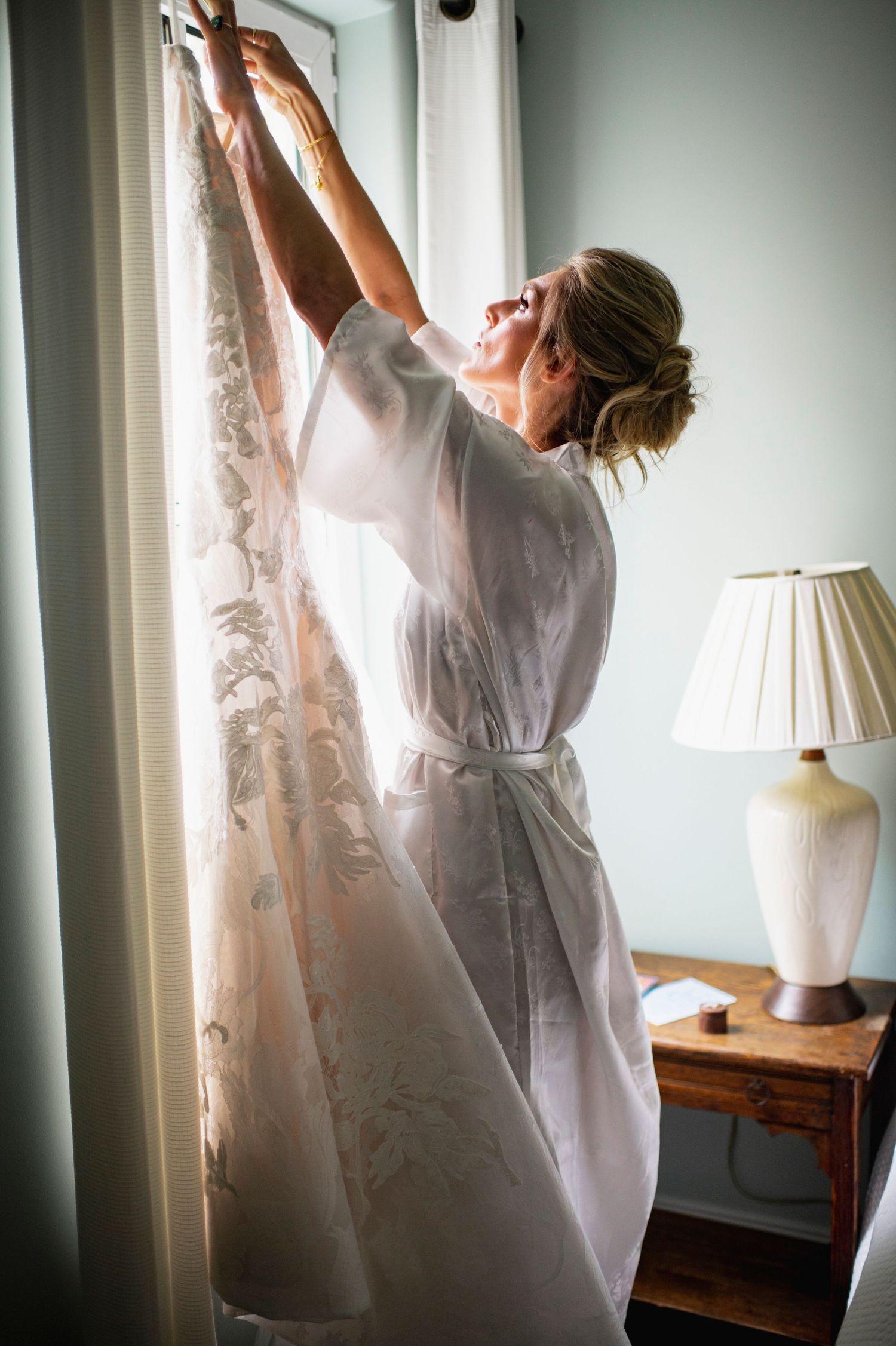 A person in a white robe reaches up to adjust curtains near a window with a table lamp in a sunlit room.