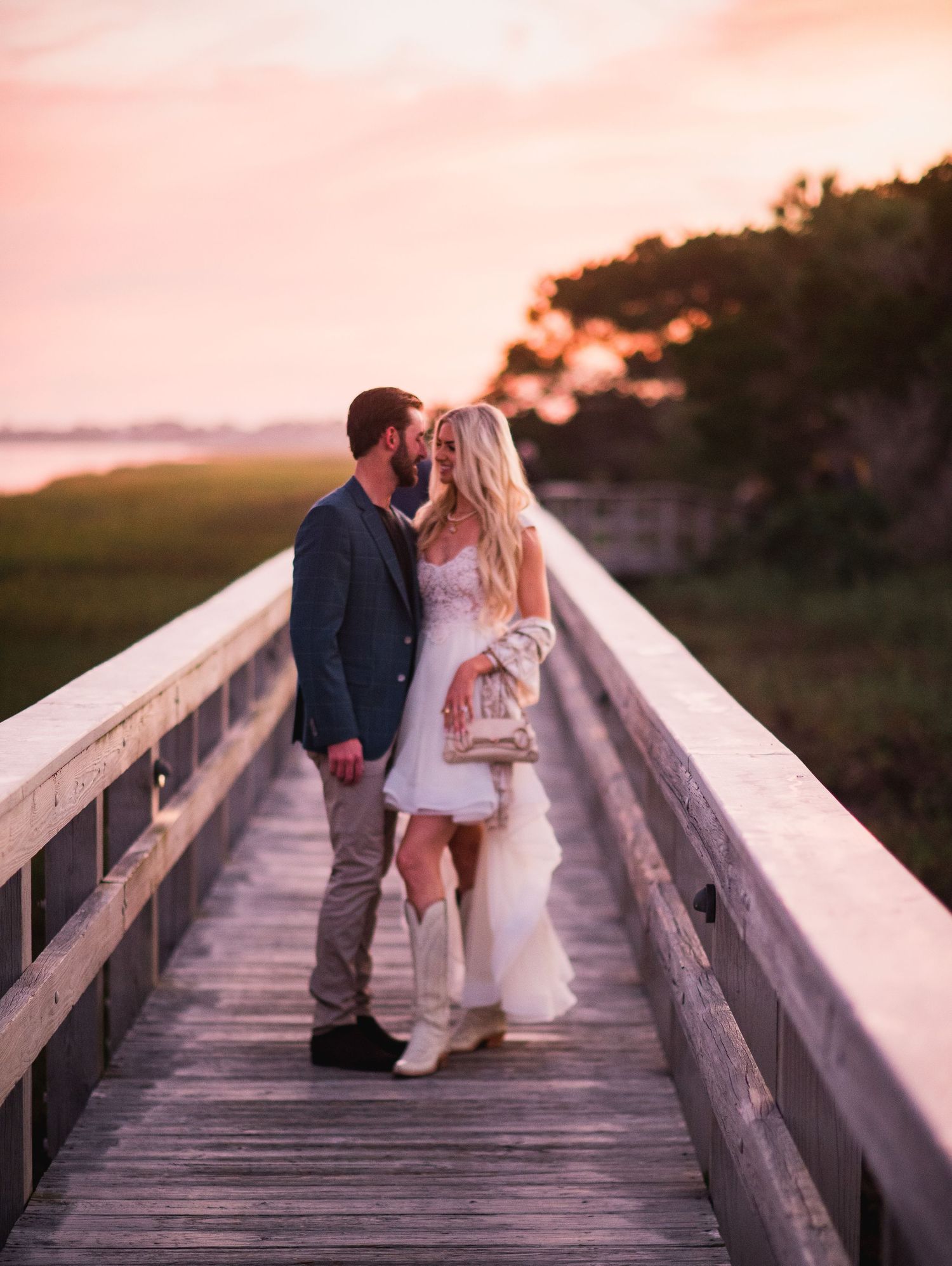 A couple shares a romantic moment at sunset on a wooden boardwalk in a coastal setting.