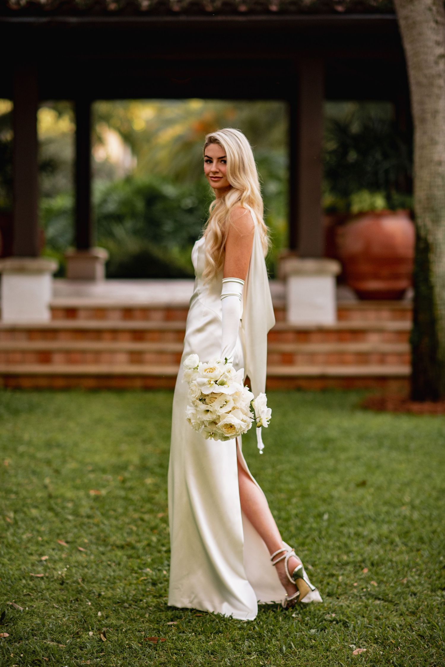 A bride in an elegant white dress holds a bouquet while walking across a lush green lawn at an outdoor venue.