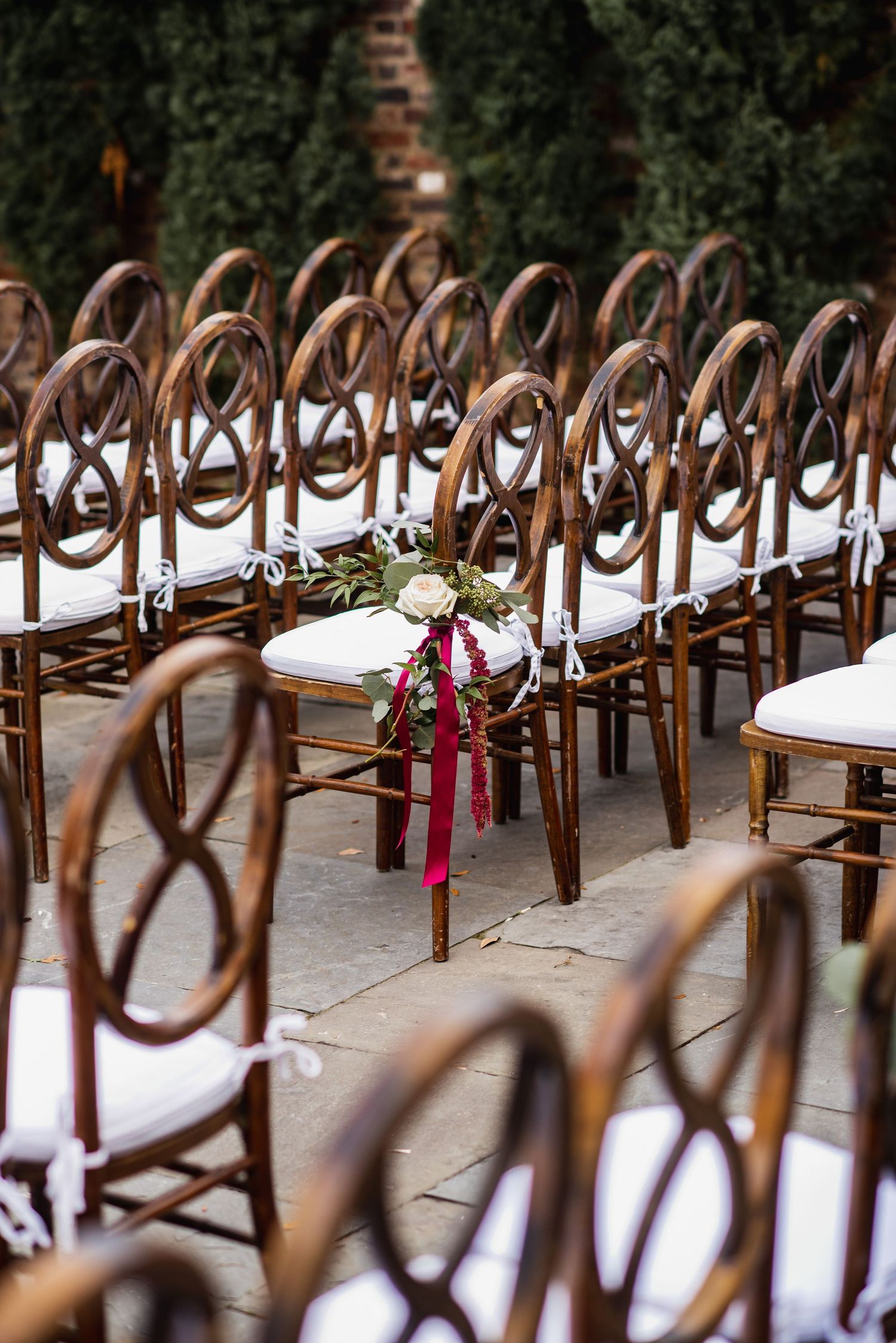 Rows of elegant wooden cross-back chairs with white cushions line up at an outdoor wedding ceremony venue.