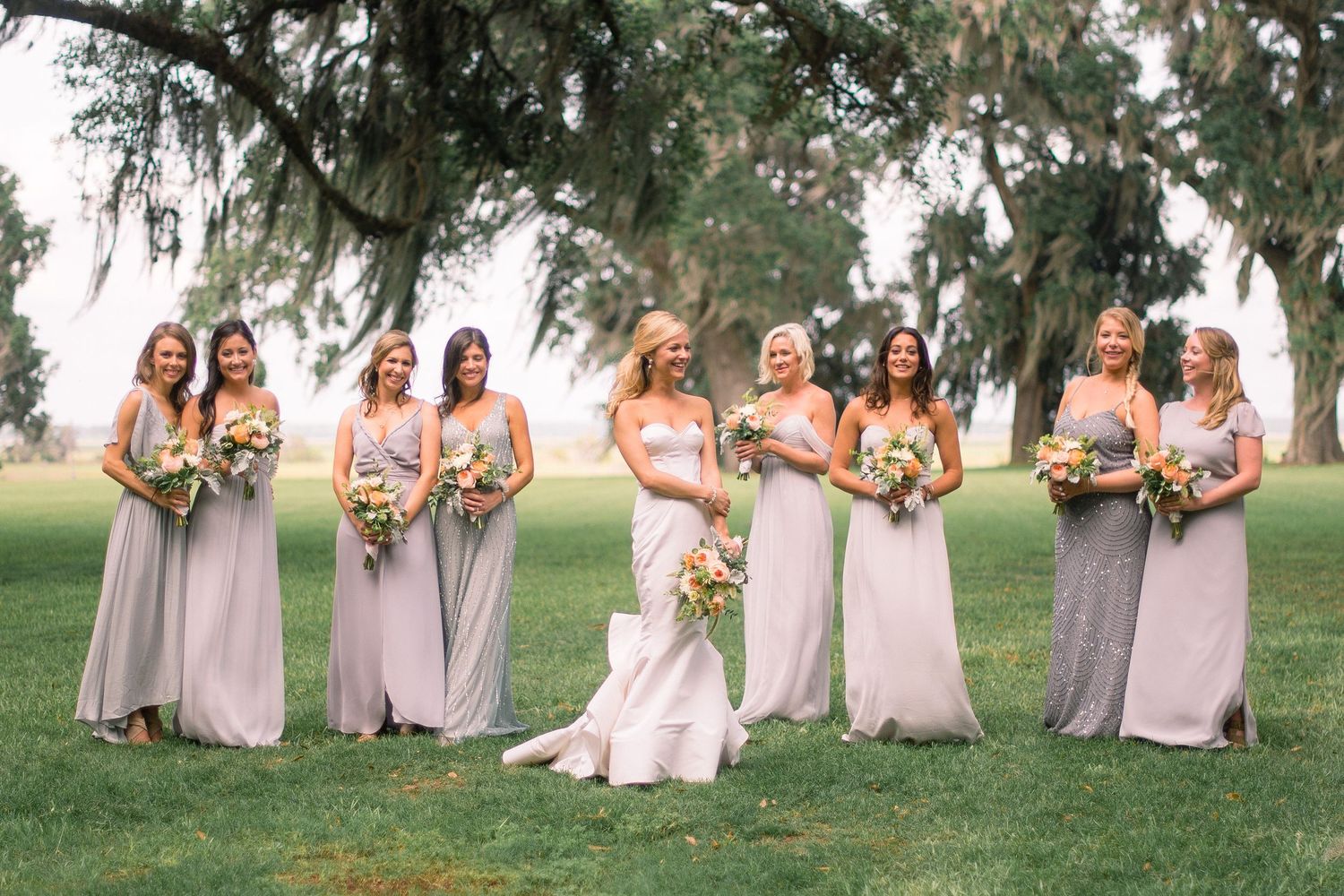 Wedding party in matching lavender dresses pose with bouquets on a grassy lawn during an outdoor celebration.
