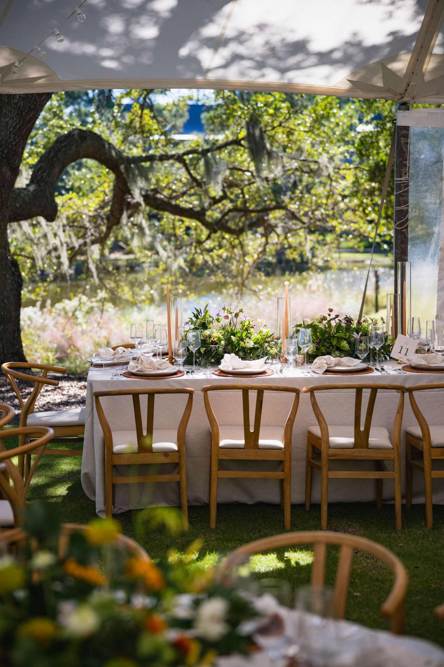 Elegant outdoor wedding reception tables set under trees with white linens and wooden chairs in dappled sunlight.