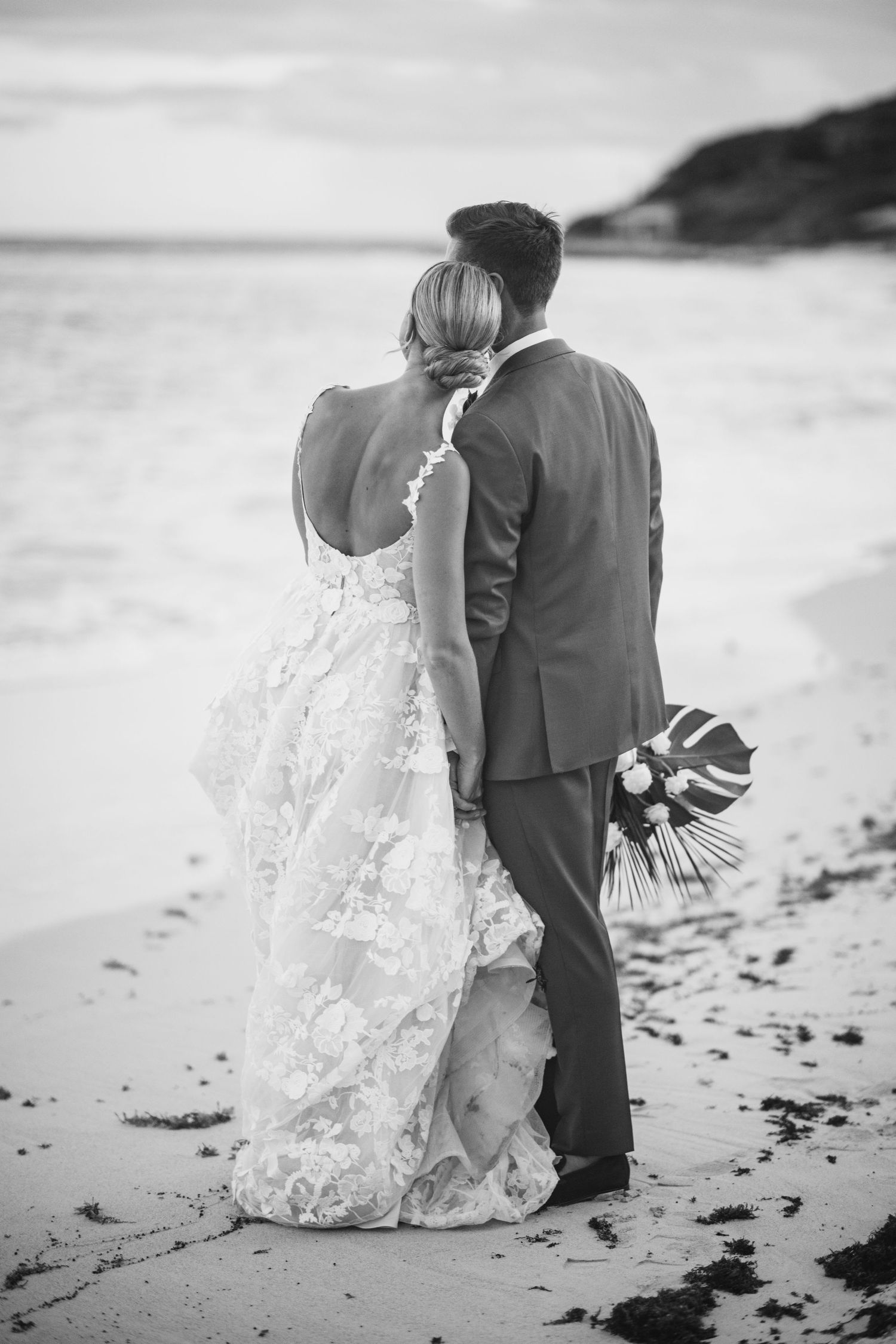 Black and white photo of couple in wedding attire walking along beach shoreline.