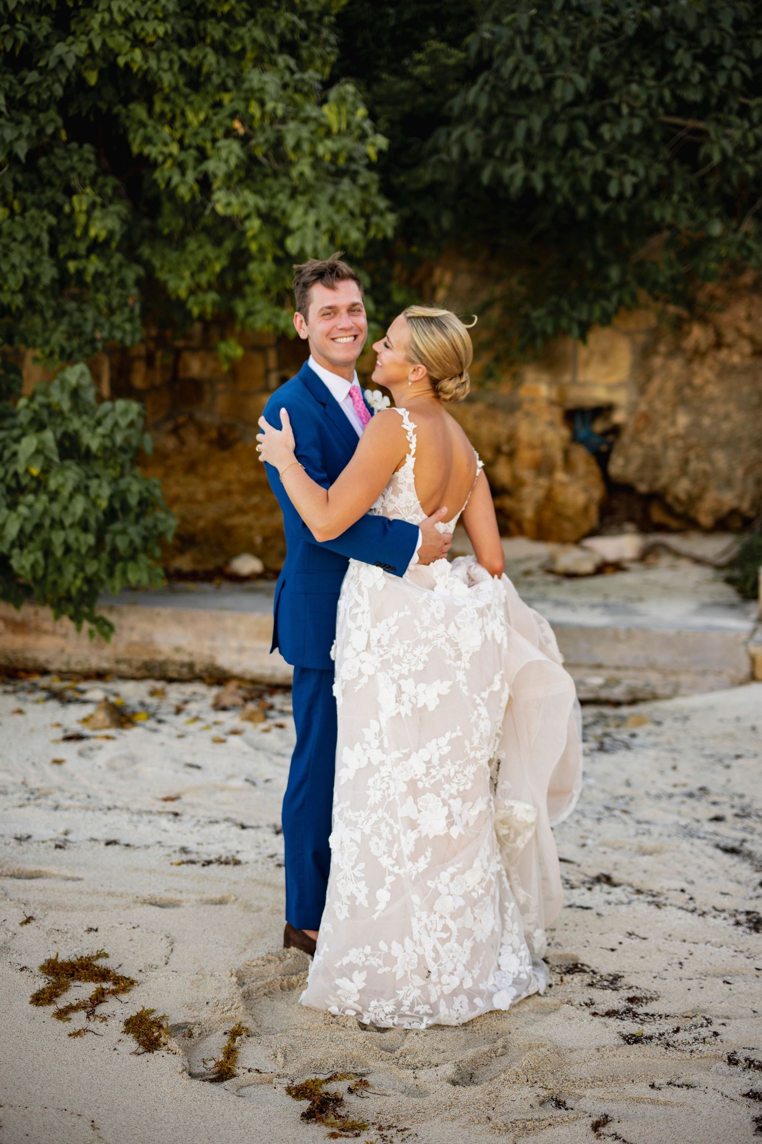 Wedding couple embracing on a scenic beach with rocky cliffs and lush greenery.