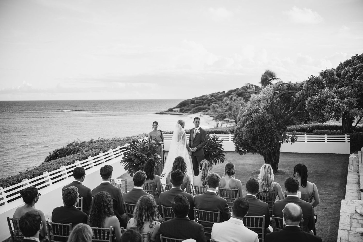 Wedding guests seated in rows watch a seaside ceremony with ocean views and coastal cliffs in the background.