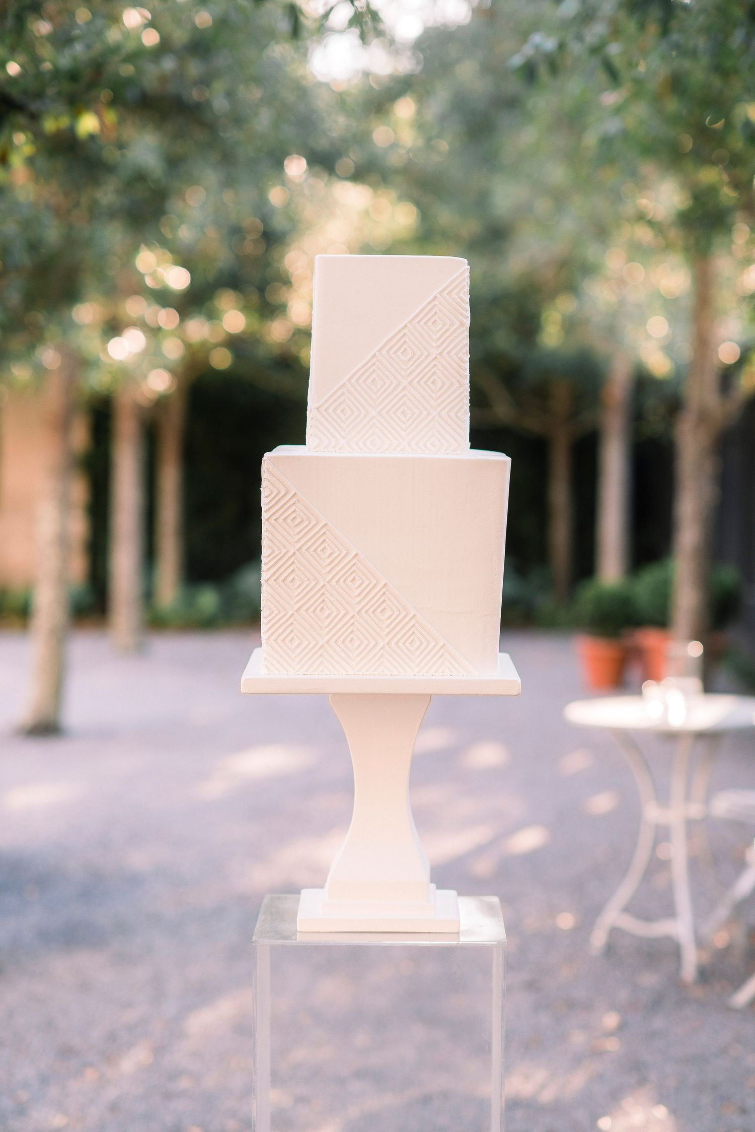 Three elegant white wedding cakes with textured details sit on display outdoors in a garden setting with trees in background.