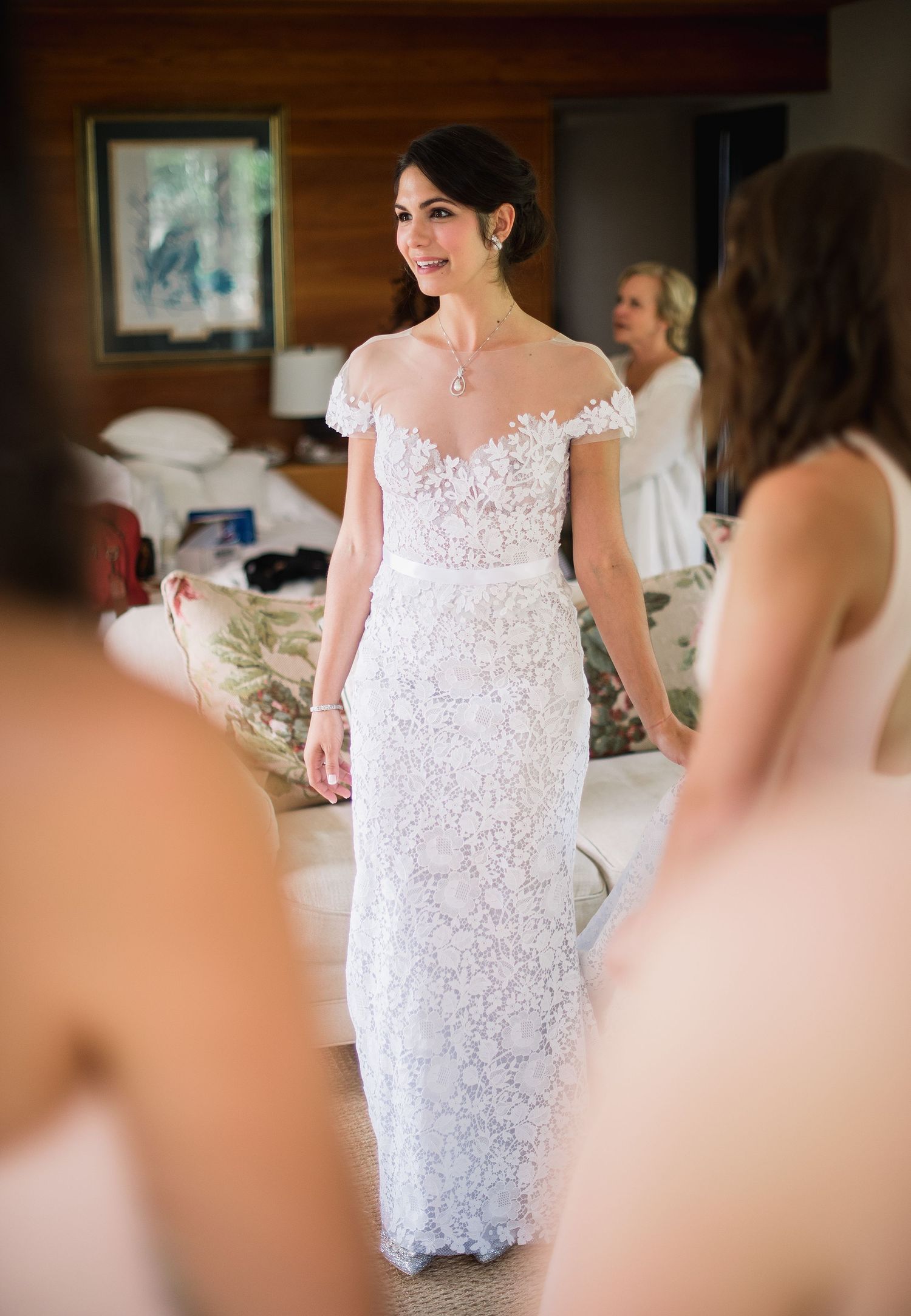 A bride wearing an elegant off-shoulder white lace wedding gown smiles while getting ready in a wood-paneled room.