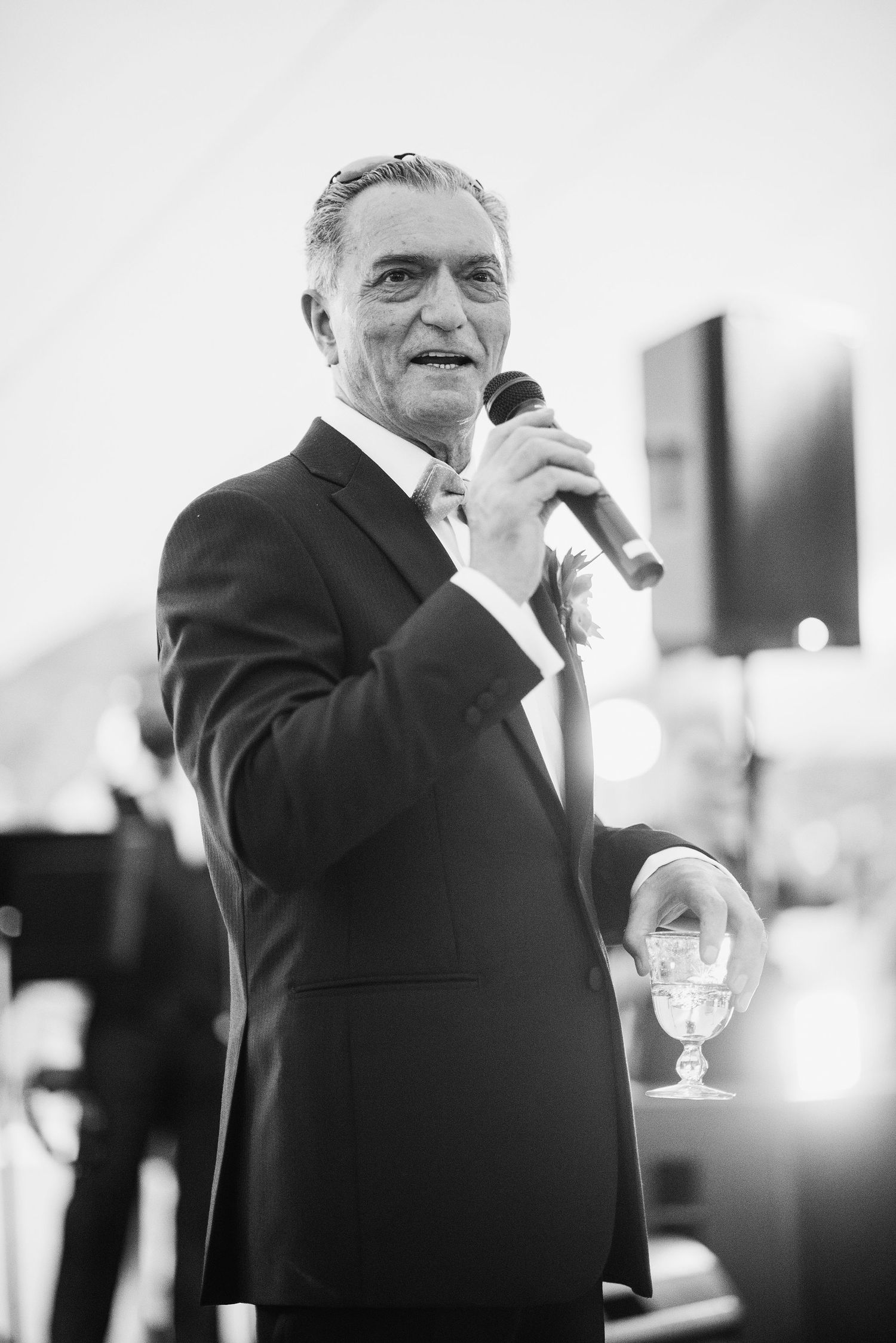 Black and white photo of a speaker in a dark suit holding a microphone and glass during a formal event.