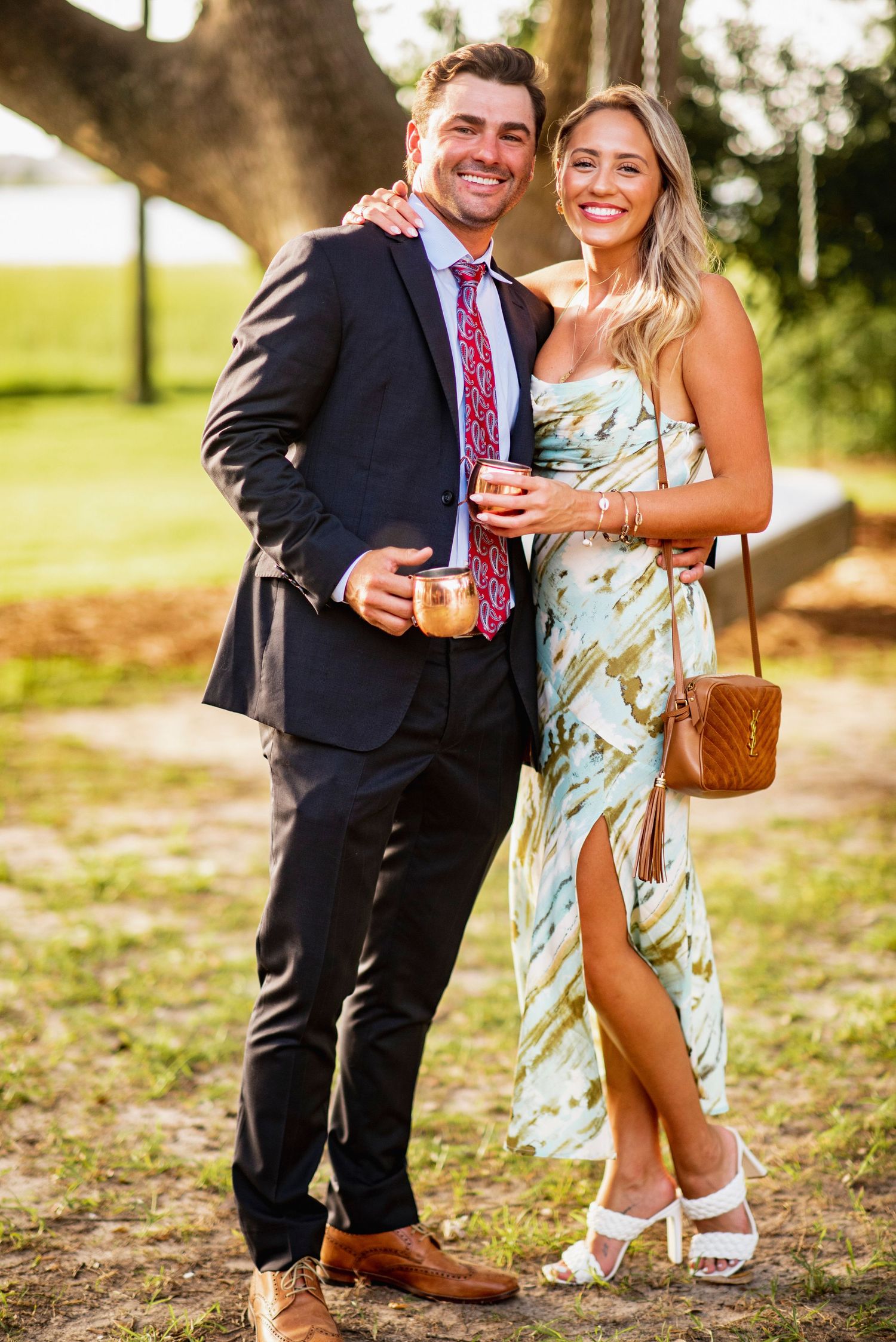 A couple dressed in formal attire share drinks at an outdoor event during golden hour sunset.