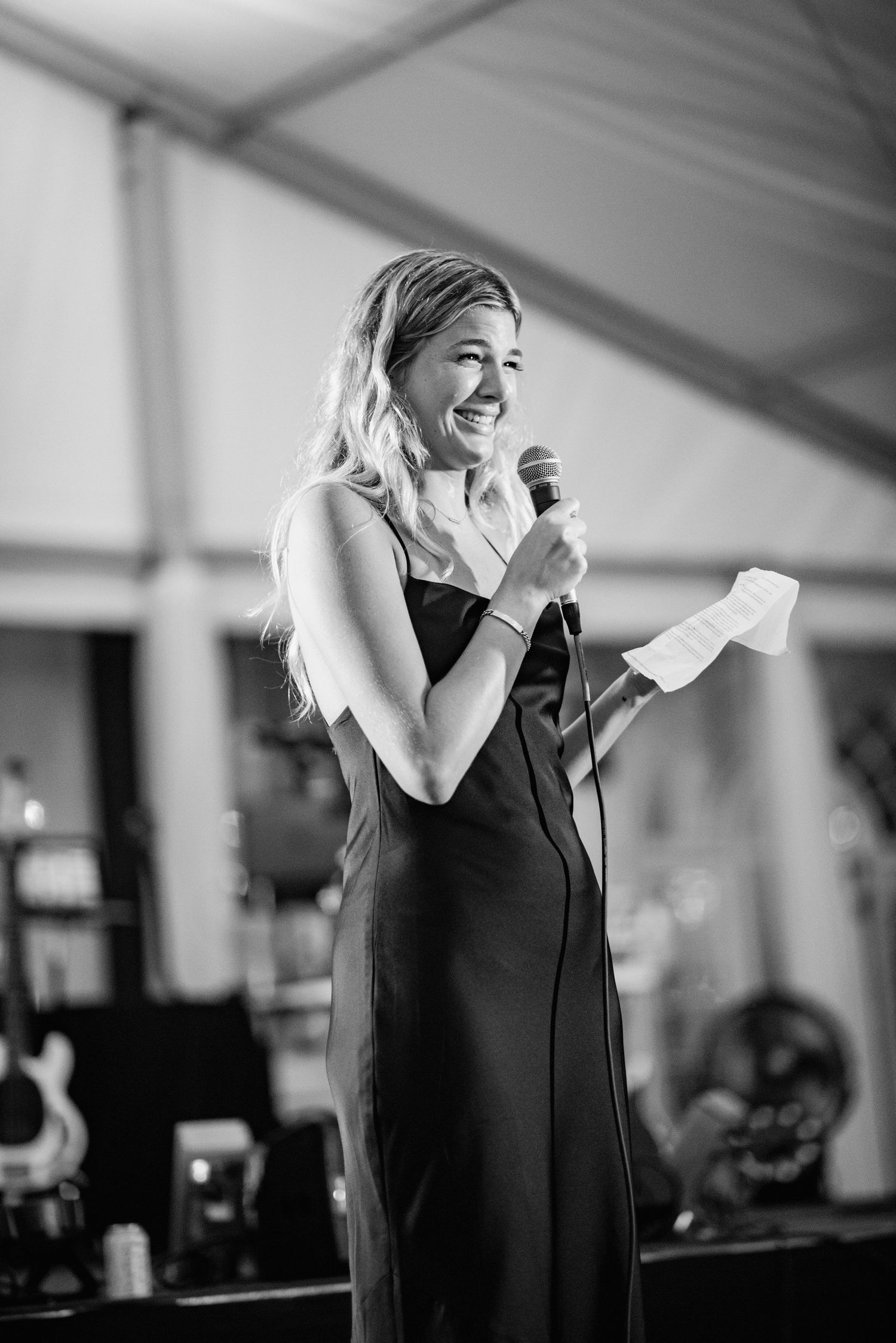 A performer in a black dress sings emotionally into a microphone during a live performance in black and white.